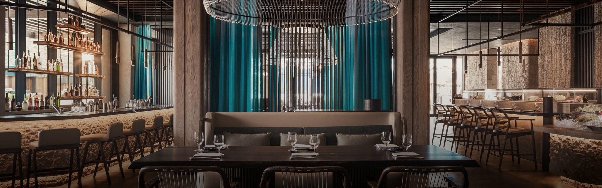 Elegant restaurant interior with a modern design, featuring large, intricate chandeliers, teal curtains, a fully stocked bar, and neatly set tables and chairs.