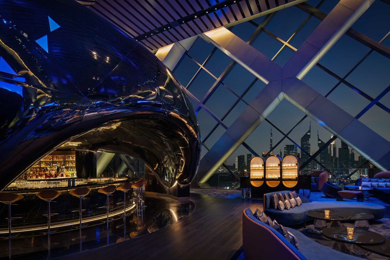 A sleek, modern rooftop bar with a curved, reflective black structure and stylish seating. Large windows showcase Dubai's skyline at dusk. Sophisticated ambiance.