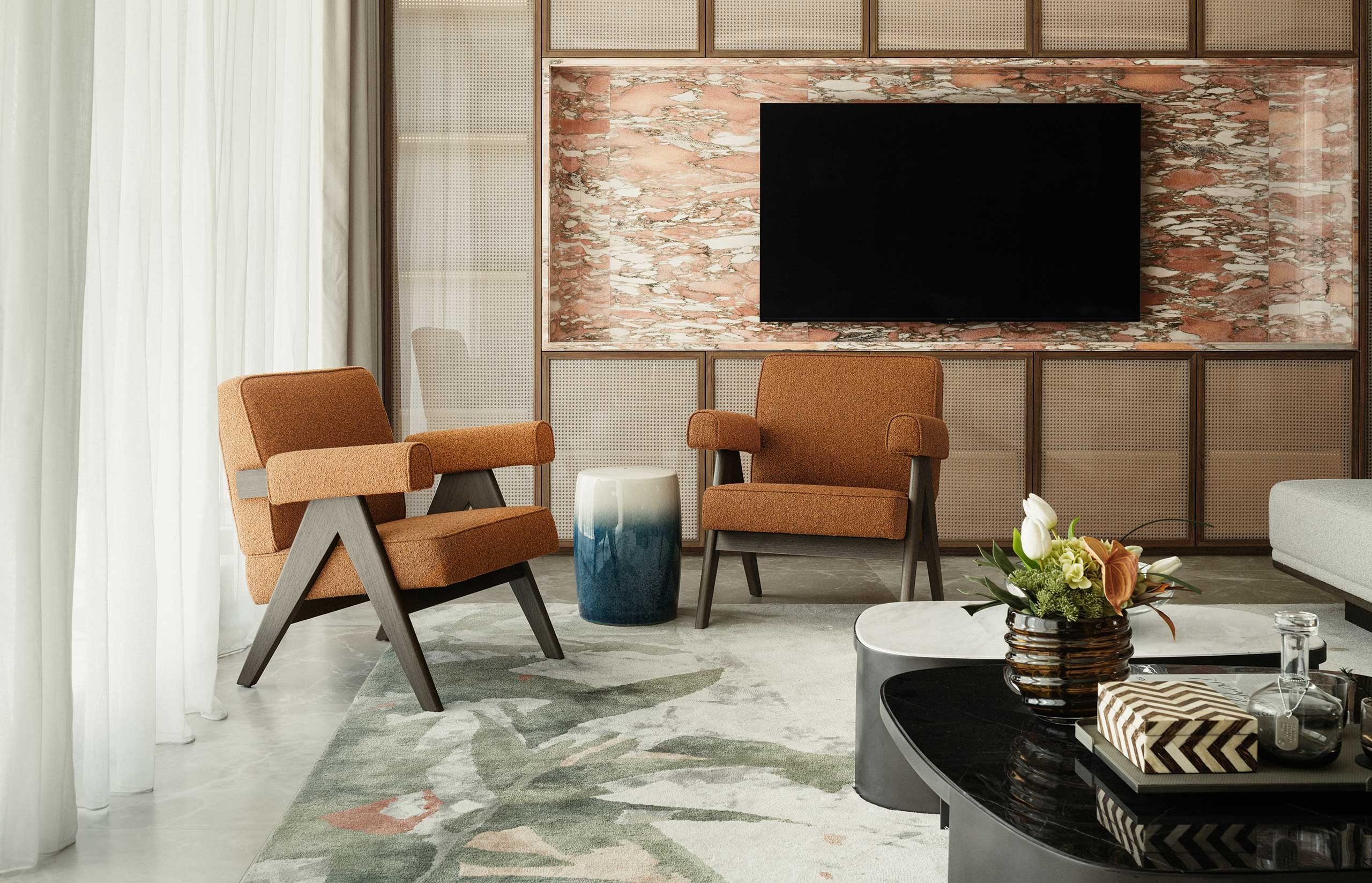 Contemporary living room with two orange armchairs, a blue and white stool, abstract rug, and large wall-mounted TV. Neutral tones evoke a calm atmosphere.
