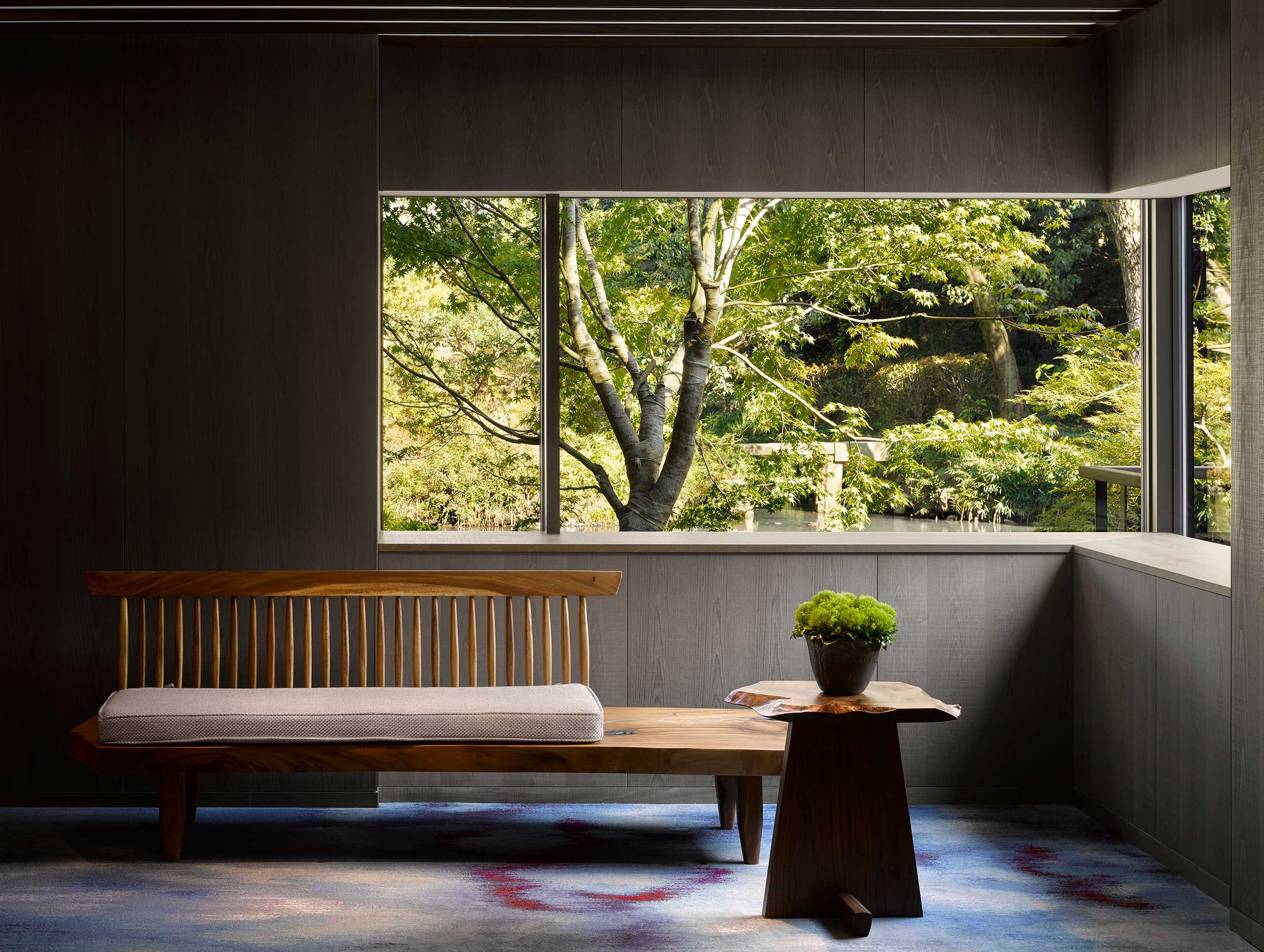 A minimalist room features a wooden bench and table with a plant, set against a large window. The view shows lush green trees, conveying tranquility.