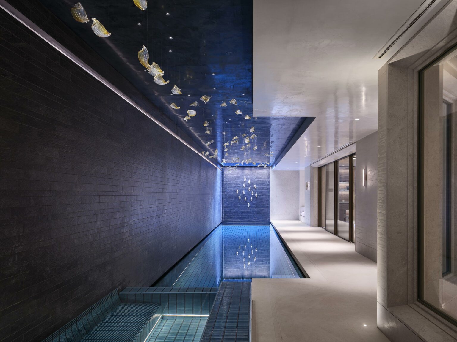 Luxurious indoor pool with a serene atmosphere. Dark walls, blue reflective water, and hanging lights create a tranquil, elegant ambiance.