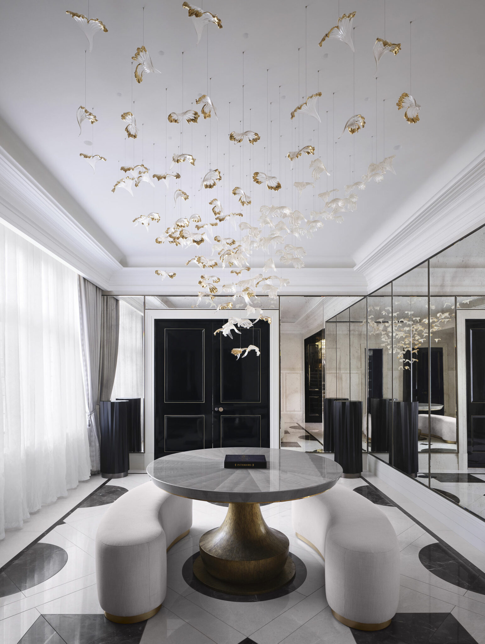 Elegant modern residential lobby with a round table and curved seating, under a chandelier of white and gold hanging ornaments. Black doors and mirrored walls add sophistication.