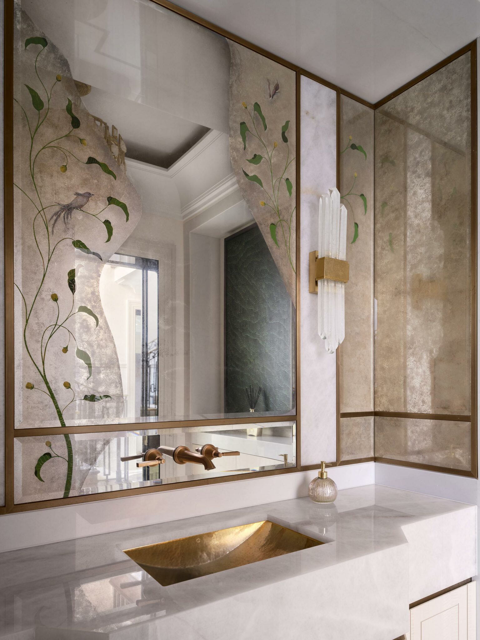 Luxurious bathroom with a gold sink set in a polished counter. Elegant wallpaper features birds and vines, with a chic vertical sconce. Reflective surfaces enhance space.