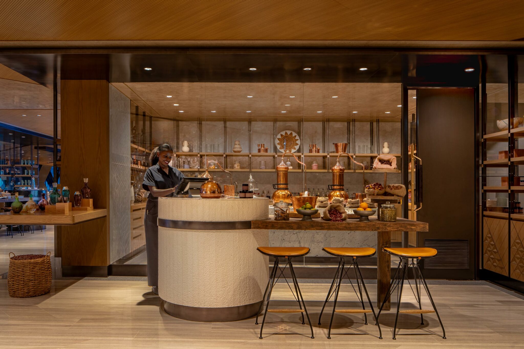 A warmly lit chef's kitchen with a waiter smiling, surrounded by copper cookware and fresh ingredients on the counter. Three stools invite guests. Cozy ambiance.
