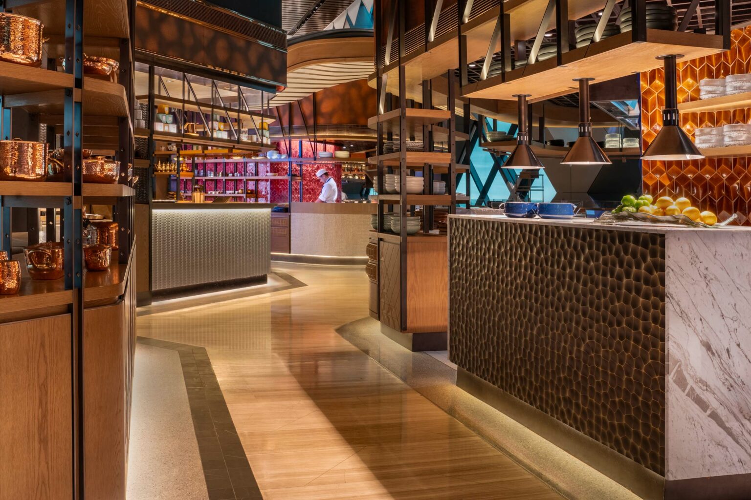 Warmly lit modern food court with wood and metal shelves, copper pots, and geometric patterns. A chef in white prepares food in the background.