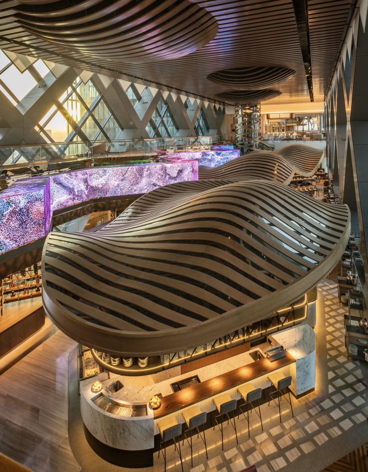 Futuristic restaurant interior with undulating, striped ceiling and colorful LED panels. Geometric window designs and a sleek bar area convey modern elegance.