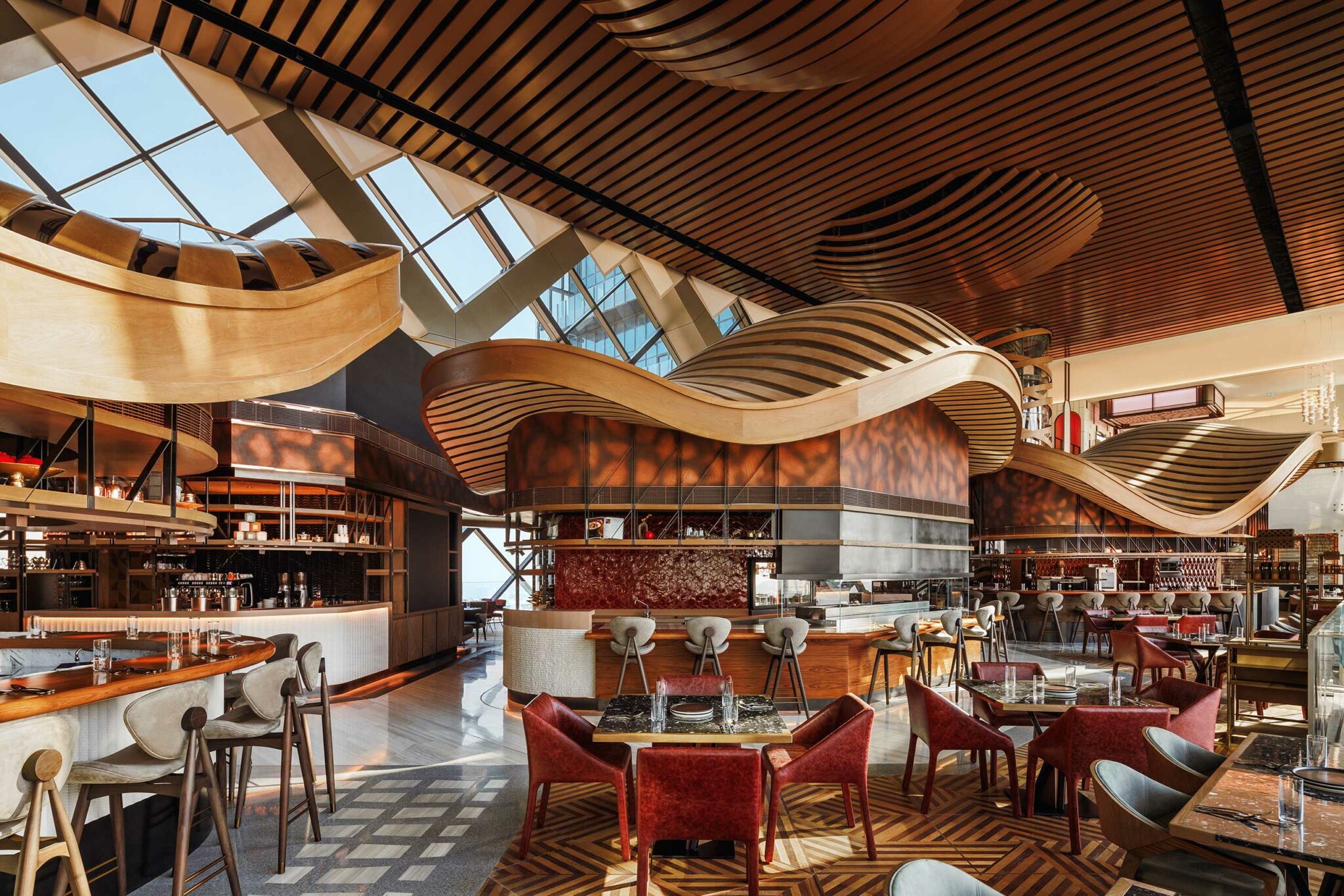 A modern food court with elegant wooden wave ceilings, geometric floor patterns, red chairs, and high bar stools. Sunlight streams through large windows.