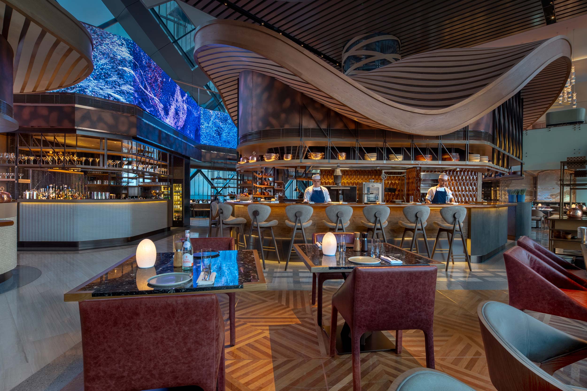 Modern restaurant interior with warm lighting, featuring a wavy wooden ceiling design, elegantly set tables, and chefs behind a sleek bar counter.