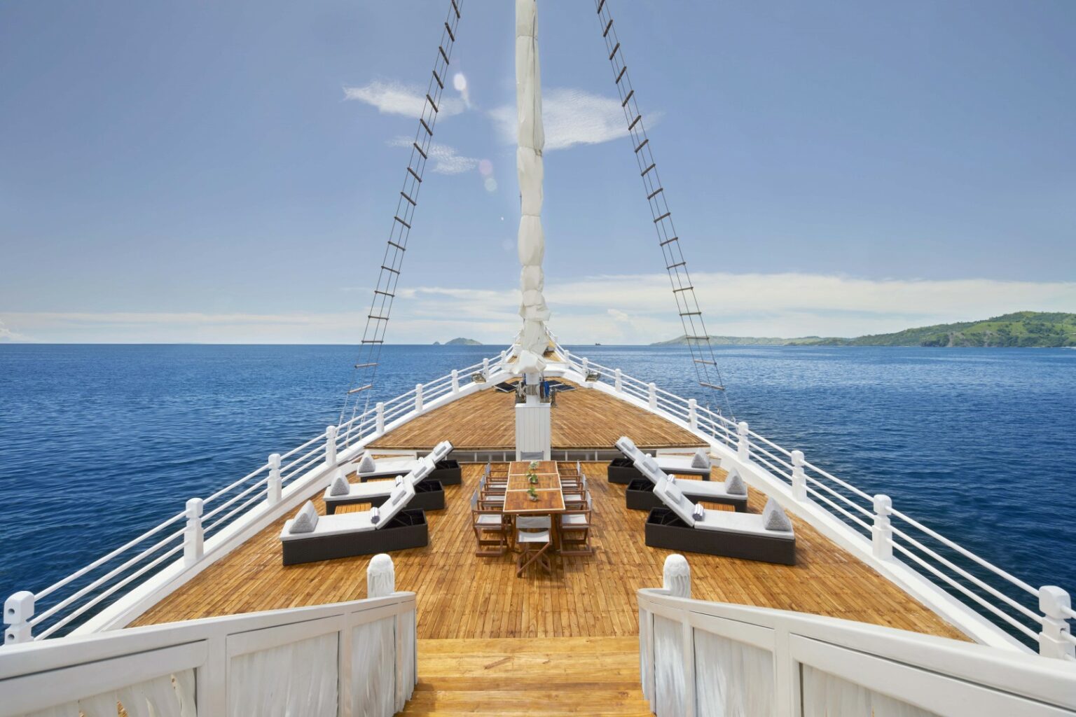 Deck of a luxurious yacht on open sea, with wooden flooring, white railings, and elegant seating areas. Clear blue sky and serene ocean in the background.