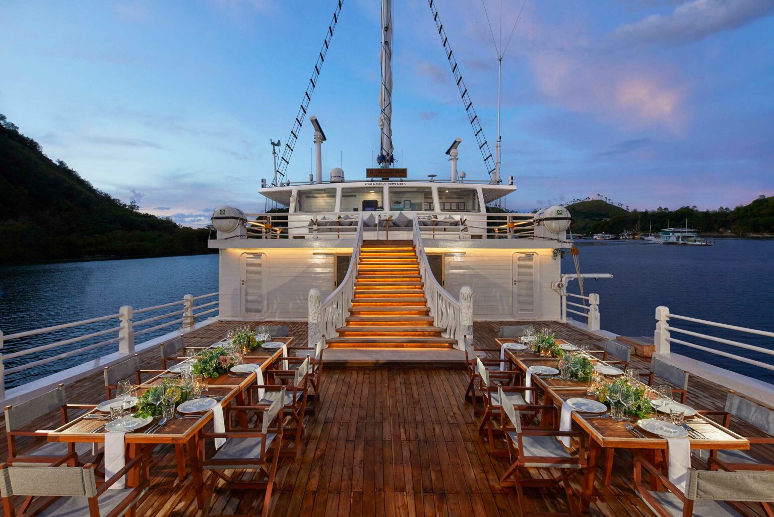 Elegant yacht deck at sunset with a grand staircase, wooden dining tables set for guests, surrounded by tranquil water and distant hills.