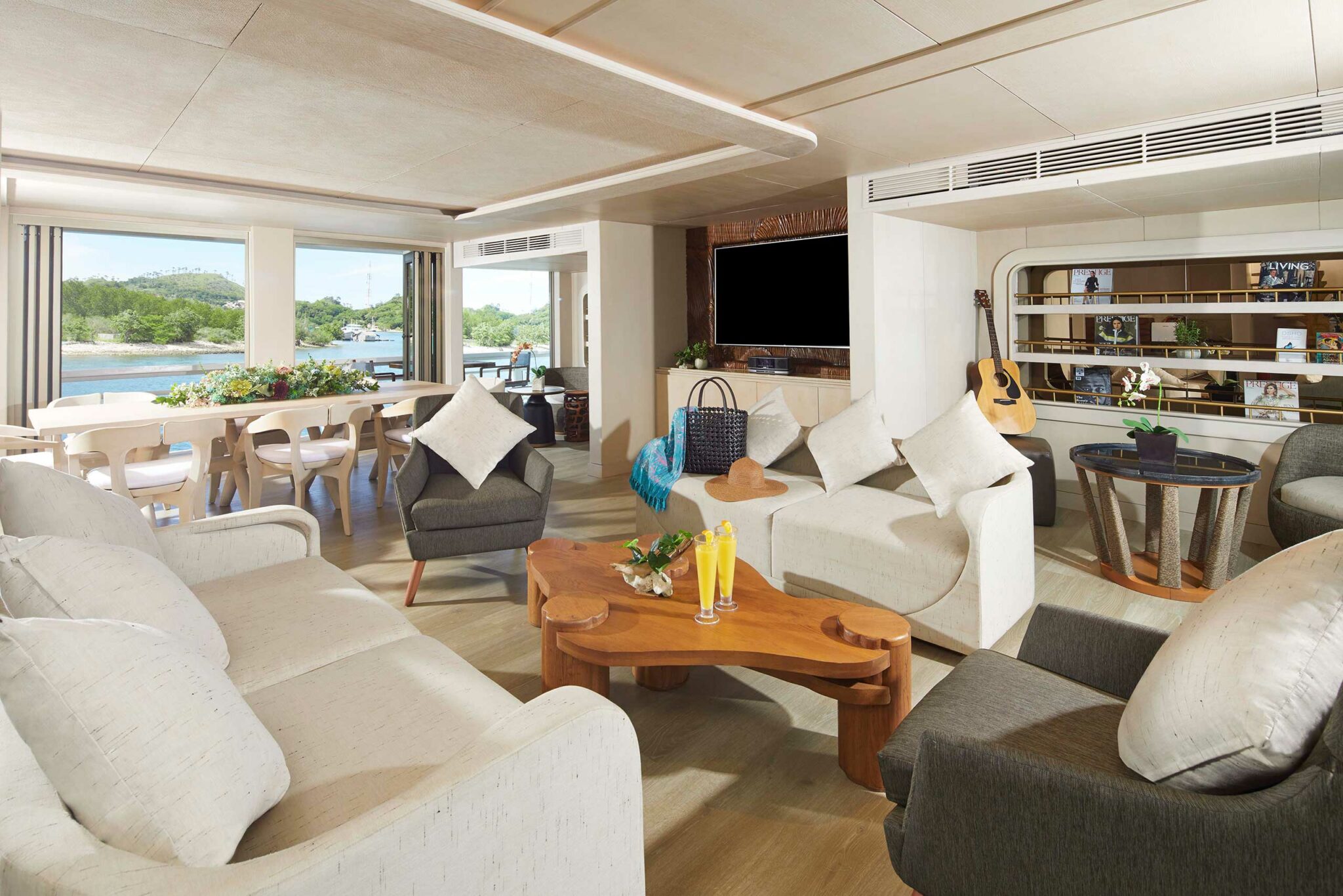 Spacious yacht living room with neutral tones, featuring white sofas, a wooden coffee table with drinks, a guitar, and a scenic view through large windows.