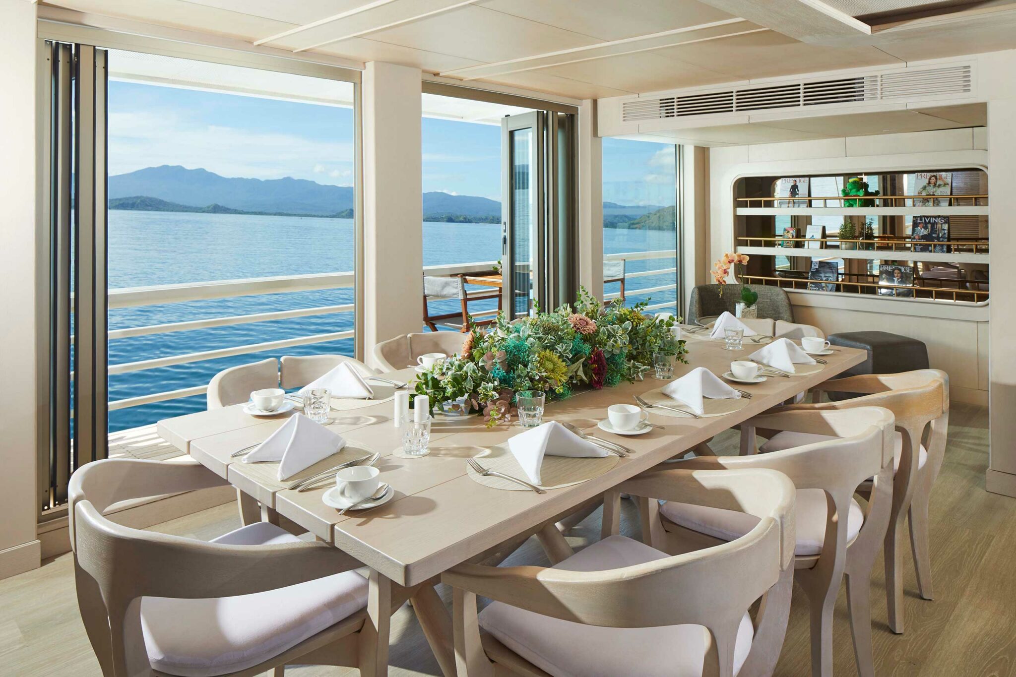 Elegant dining room on a yacht with a view of the sea and mountains. Large table set with white napkins, cups, and a colorful floral centerpiece.