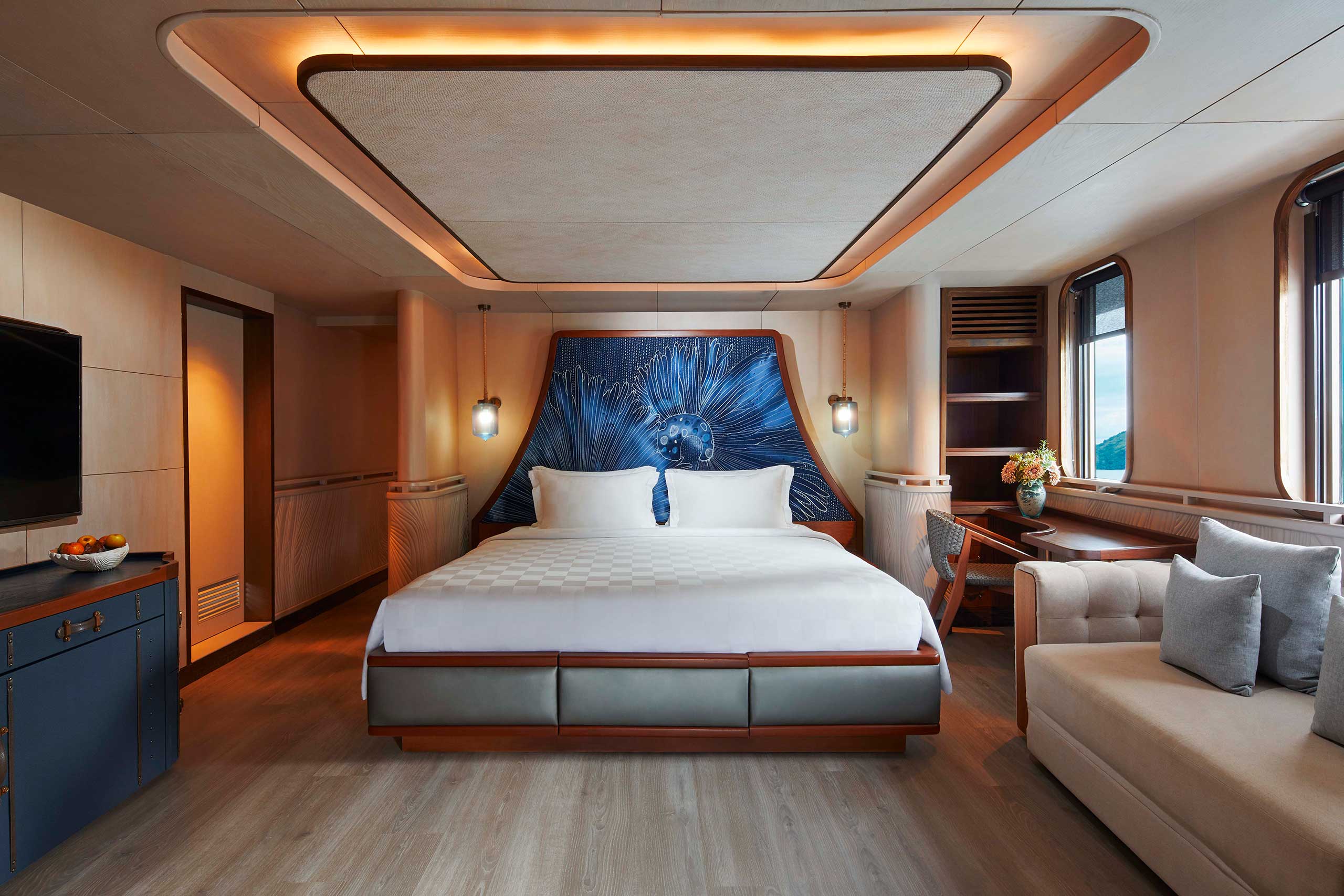 Luxurious yacht cabin with a king-sized bed, white linens, and a vibrant blue abstract headboard. Soft lighting and modern decor create a serene atmosphere.