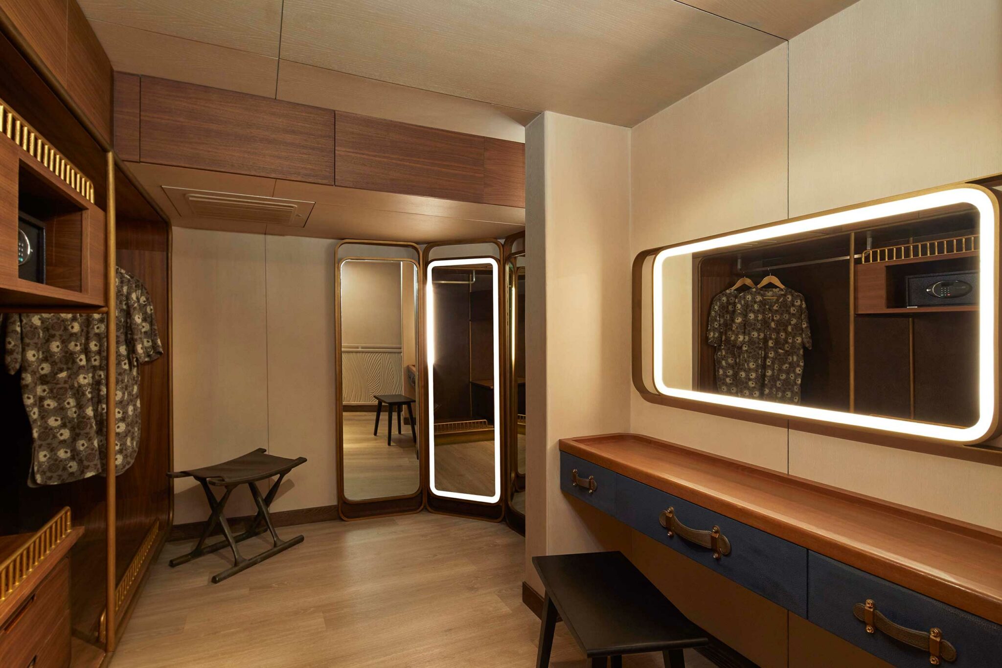 Modern yacht dressing room with a wooden wardrobe, illuminated mirrors, and a blue vanity. A stool and patterned shirt add a cozy, stylish touch.