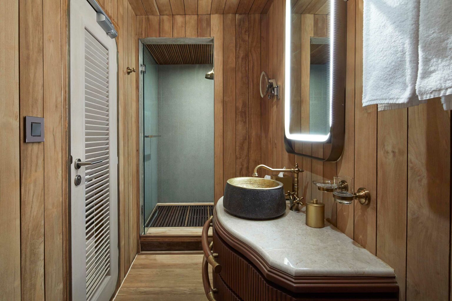 Elegant yacht bathroom with wooden walls features a stone basin on a marble counter, brass fixtures, an illuminated mirror, and a modern glass shower. Warm ambiance.