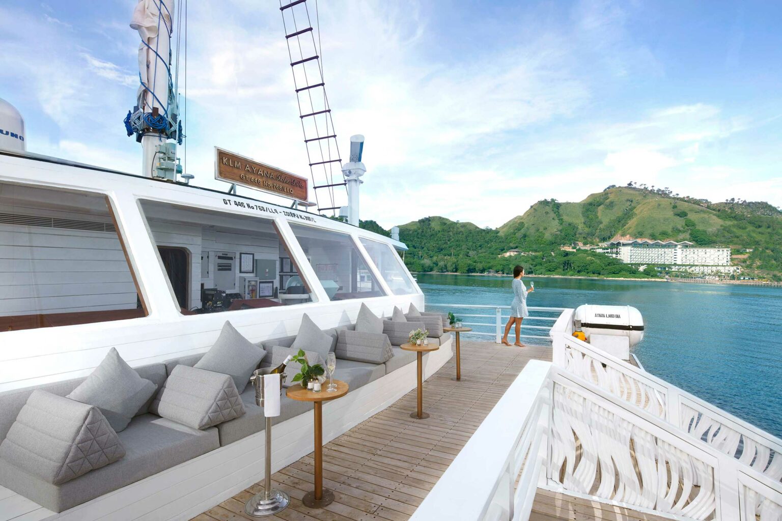 Luxurious yacht deck with plush seating and tables arranged along a white cabin. A person stands gazing at serene water and lush green hills.