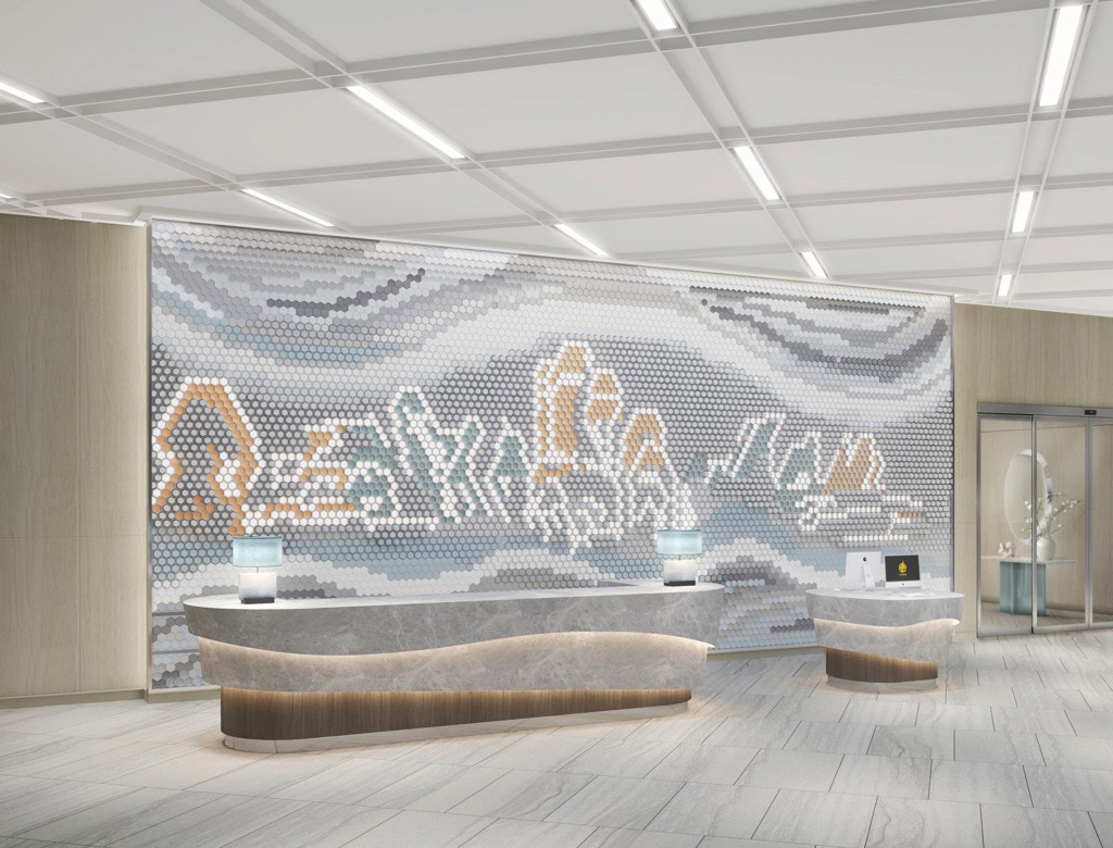 Modern hotel reception area with a sleek, curved desk and two lamps. The backdrop features a large, artistic mosaic wall with abstract patterns in soft, neutral tones.