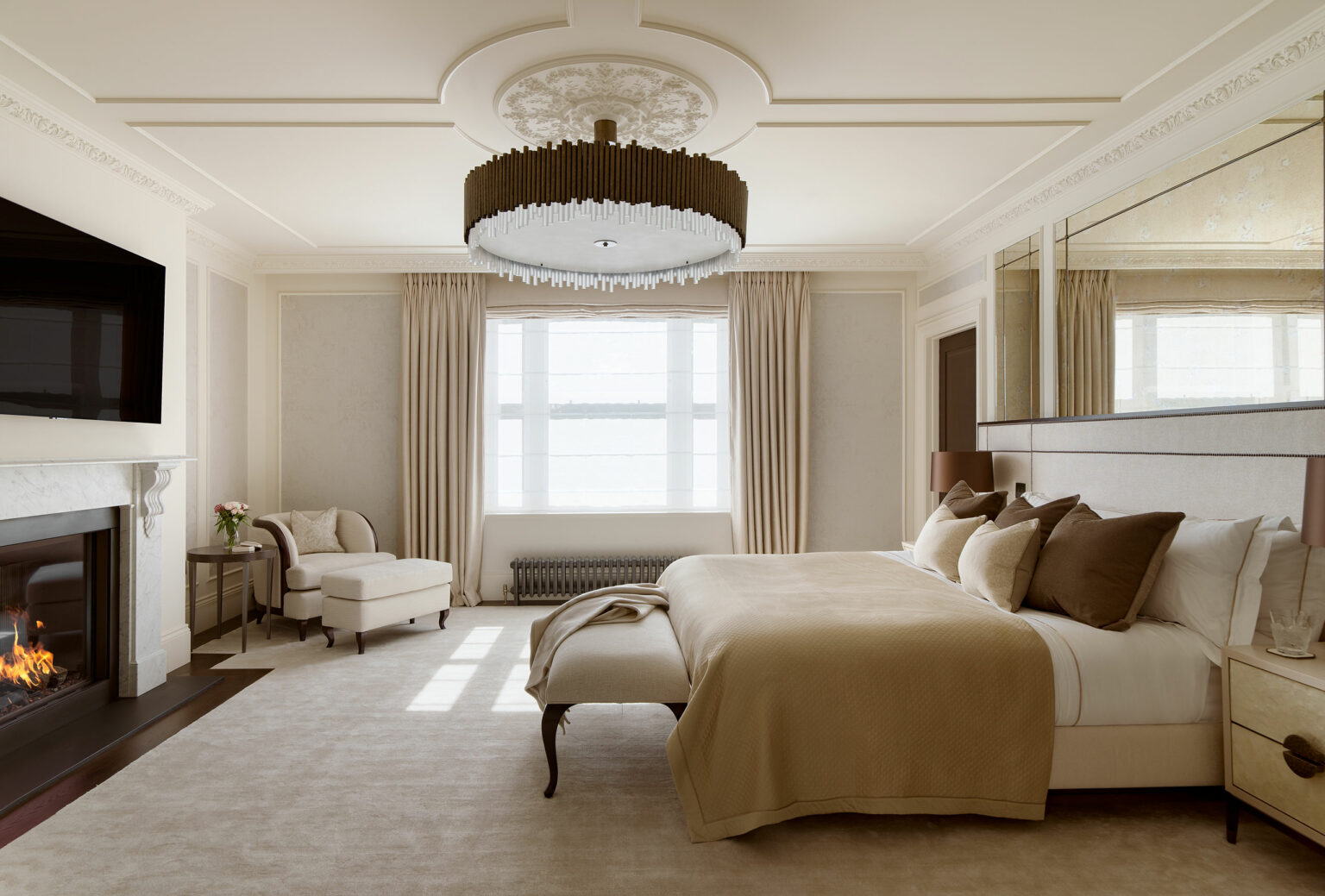 Elegant bedroom with beige tones; a plush bed with cushions, a stylish chandelier, large windows with curtains, and a cozy fireplace on the left.