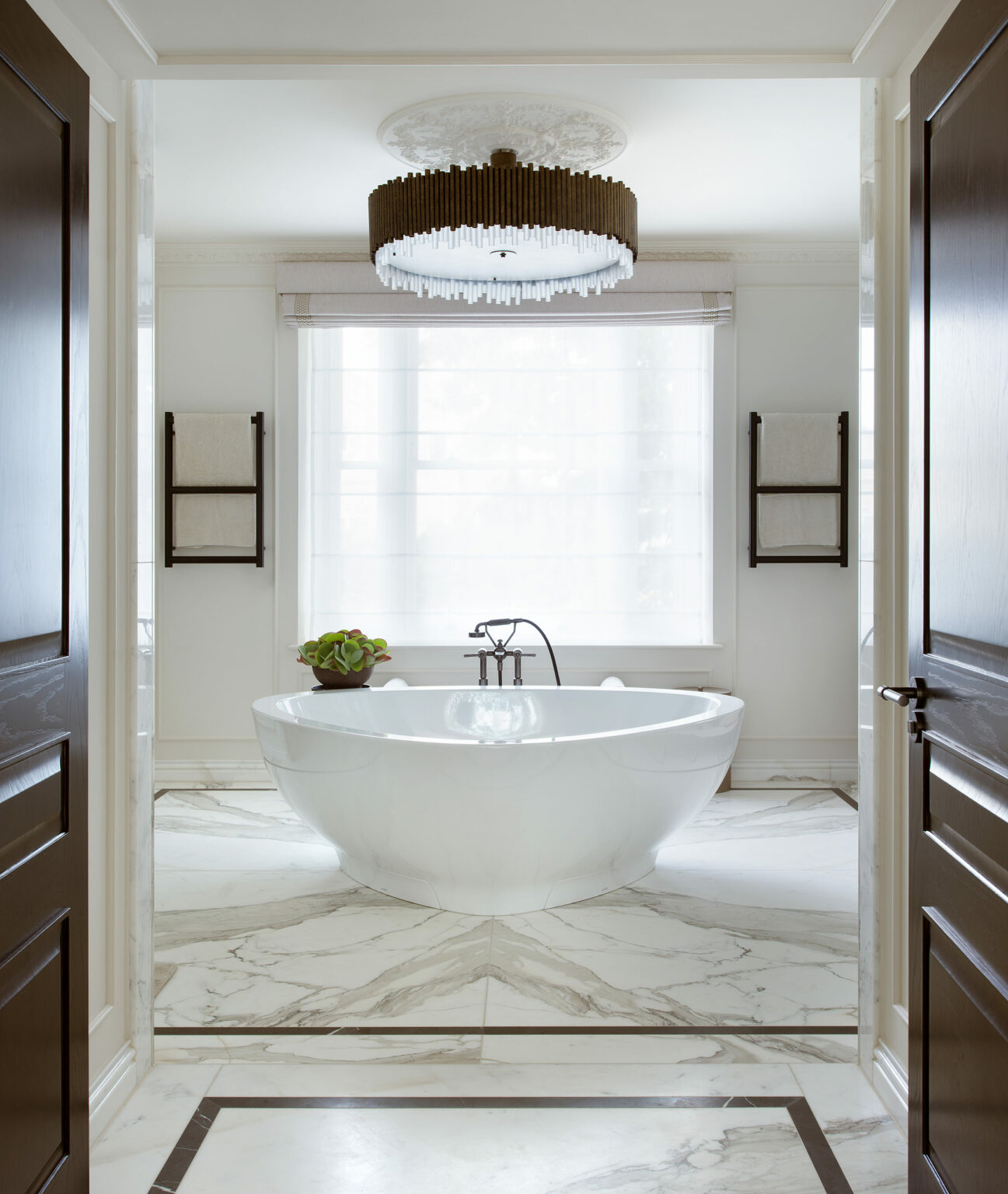 Luxurious bathroom with a freestanding white tub centered on marble floors. Large window with sheer curtains, elegant chandelier, and towel racks create a serene ambiance.