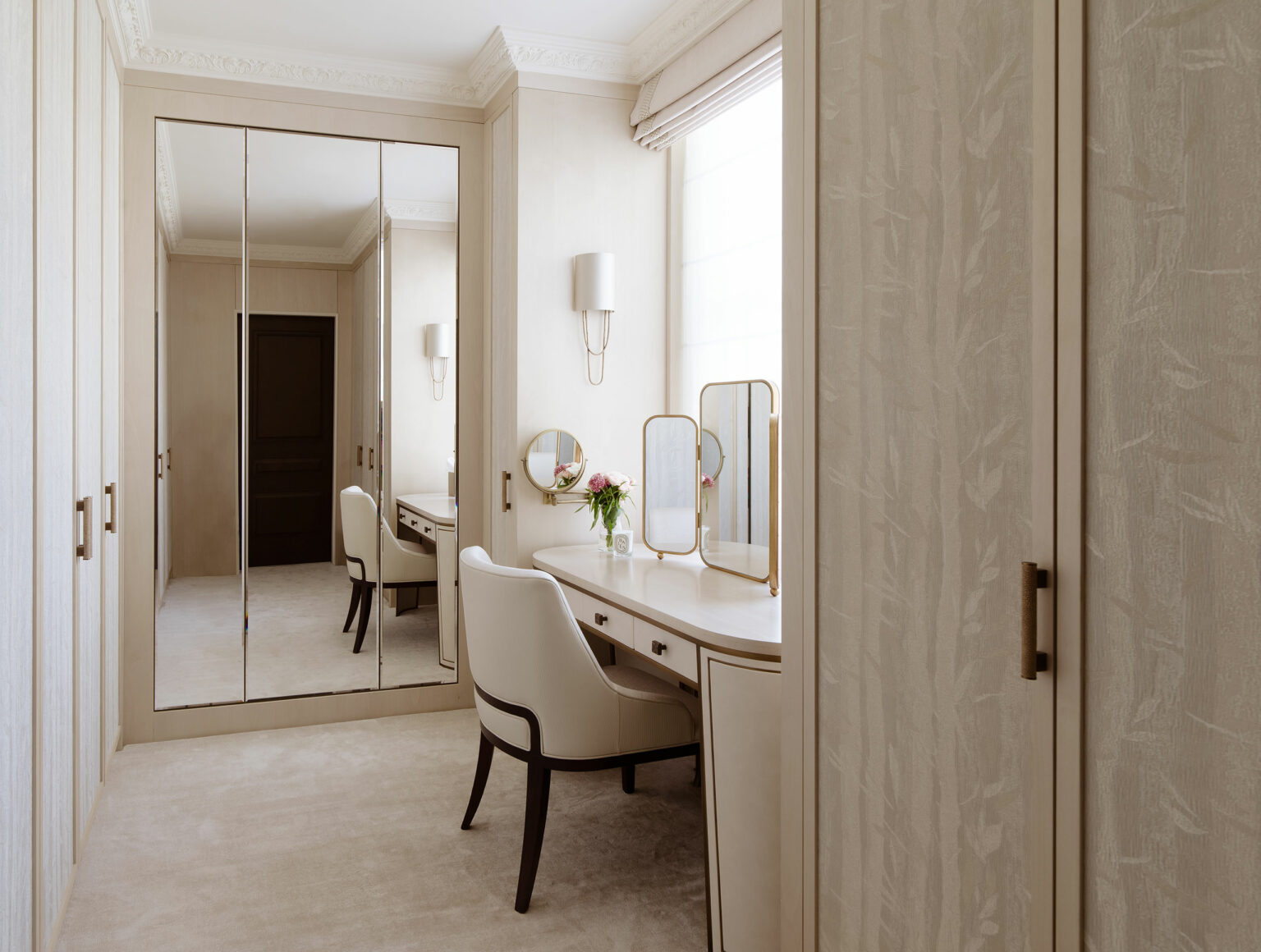 Elegant dressing room with a light beige color palette. Features mirrored wardrobe doors, a vanity table with fresh flowers, and a comfortable white chair.