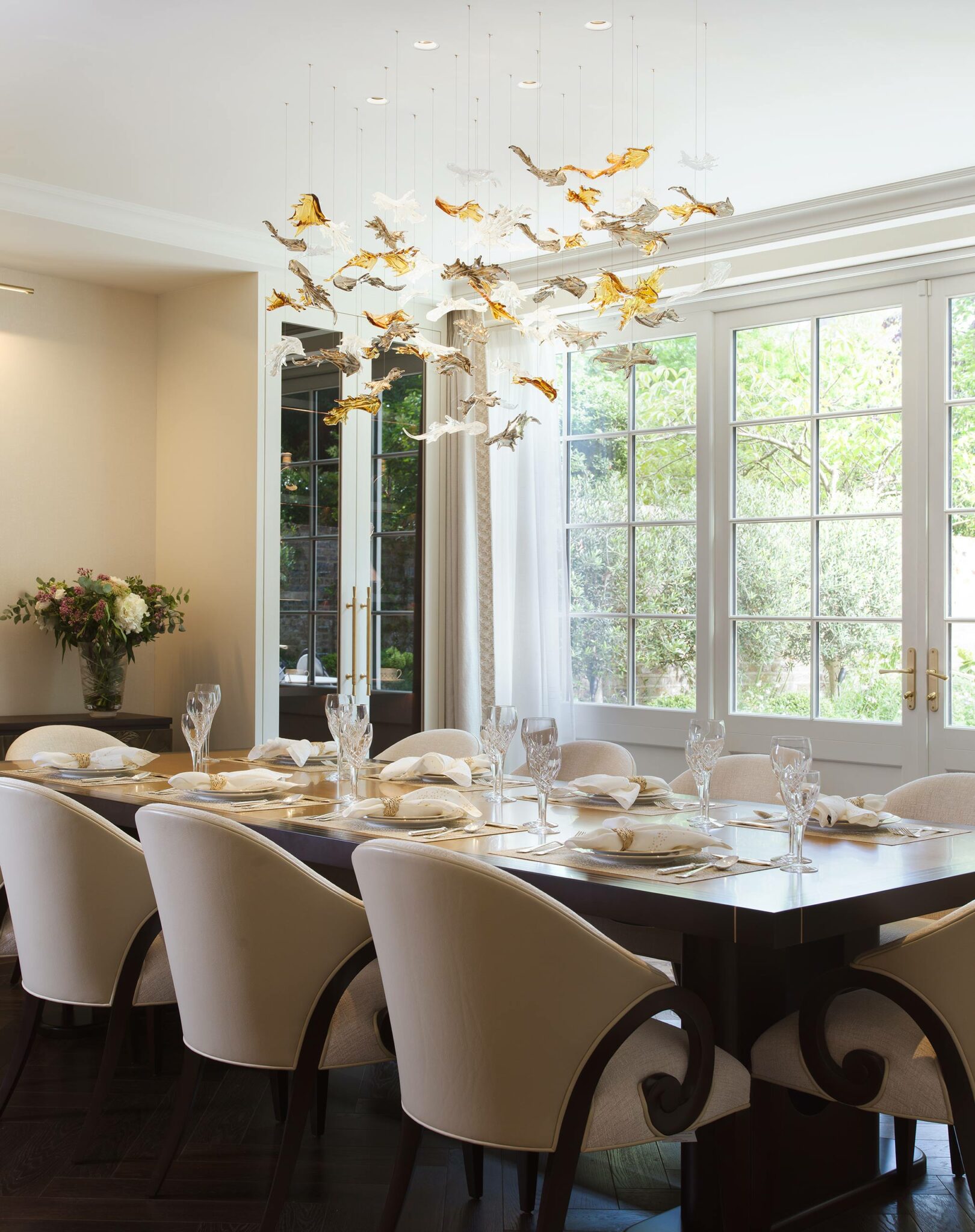 Luxurious dining room featuring a polished table set for eight with white chairs. Elegant glass bird chandelier and large windows with garden views enhance the serene atmosphere.