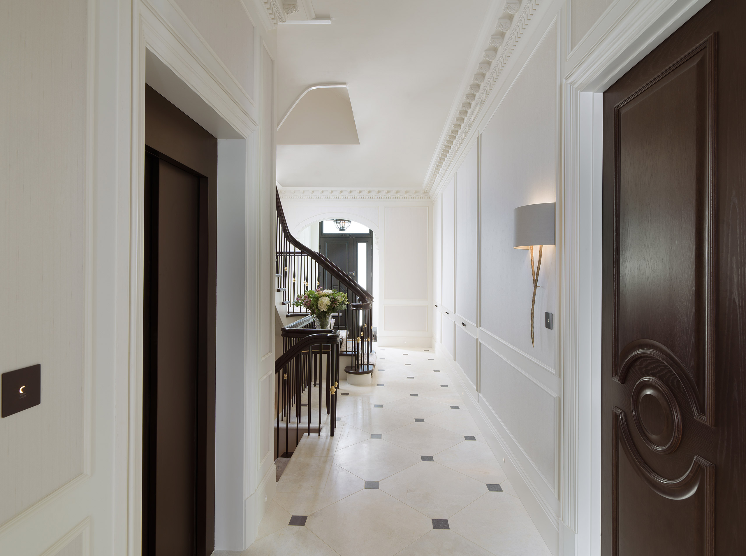 Luxurious hallway with elegant wooden staircase, vase of flowers on a console table, white panel walls, dark wooden door, and wall-mounted lamp.