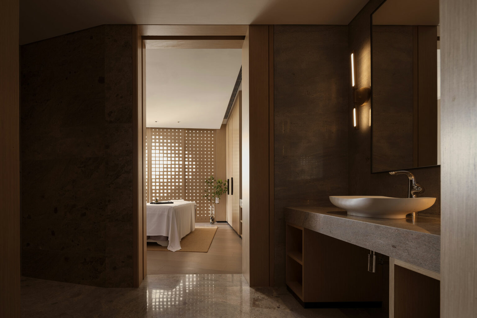 Modern spa interior of a luxury hotel with dim lighting. Foreground has a stone sink counter, foreground place leads to a softly lit massage room with a treatment table, wooden accents, and a potted plant. Calm, serene ambiance.