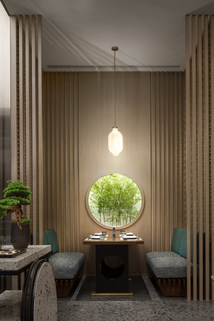 A cozy dining nook featuring a round mirror reflecting greenery, surrounded by stylish booths. Soft lighting from a pendant lamp enhances the inviting atmosphere, making it perfect for intimate dining in a contemporary restaurant setting.