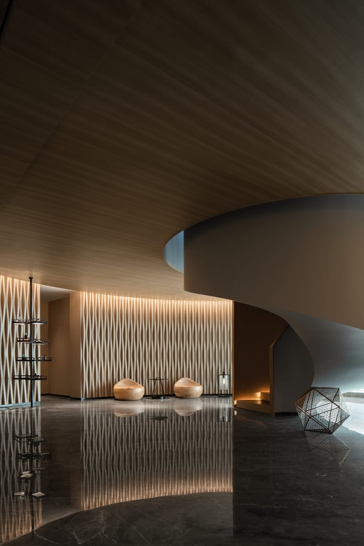 A sleek spa lobby featuring a modern spiral staircase, stylish seating, and textured wall panels, with soft lighting reflecting on polished floors, creating a sophisticated and inviting atmosphere.