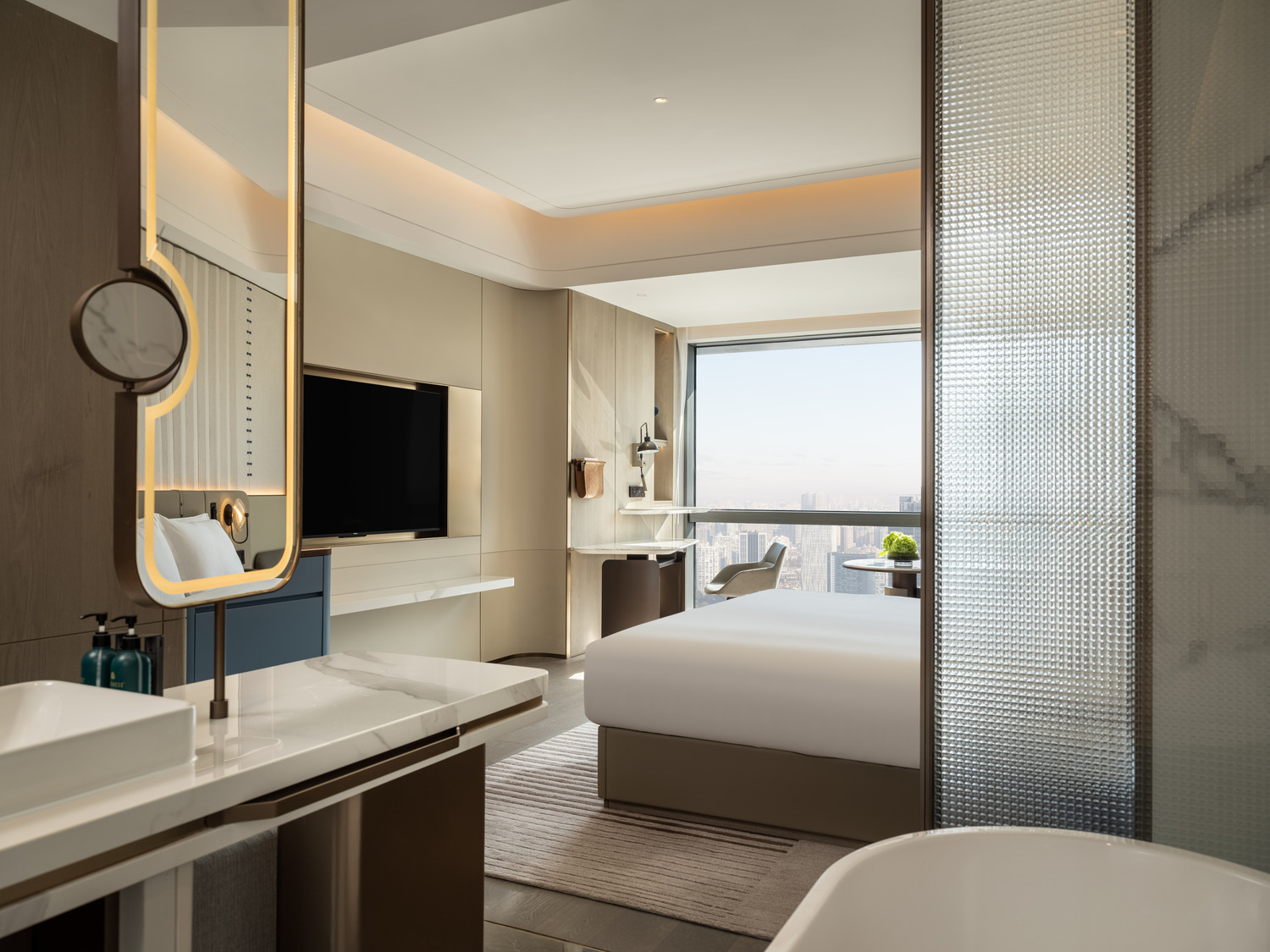 Modern hotel room with neutral tones featuring a large bed, marble bathroom with a freestanding tub, sleek furniture, and soft lighting, conveying relaxation.