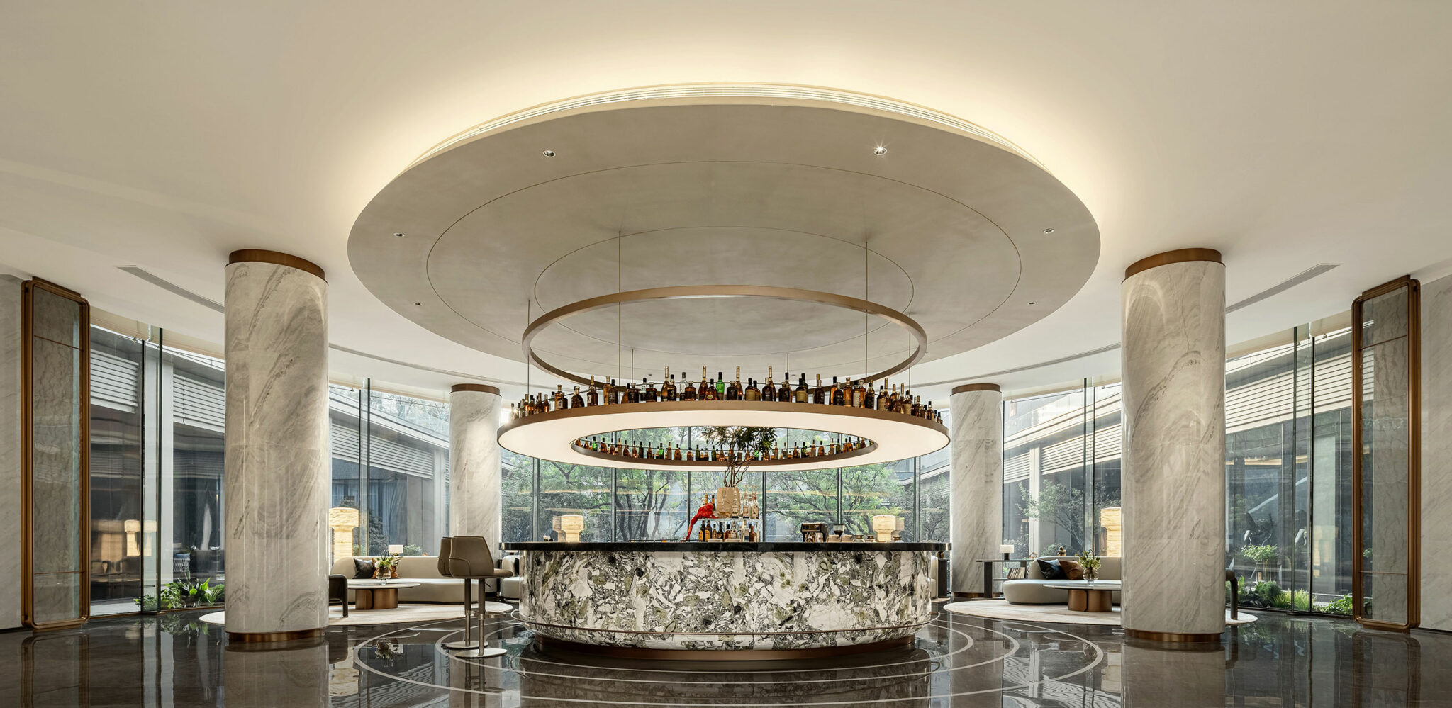 Luxurious hotel lobby bar with a circular marble counter, suspended liquor shelves, and elegant lighting. Large windows and modern decor create a sophisticated ambiance.