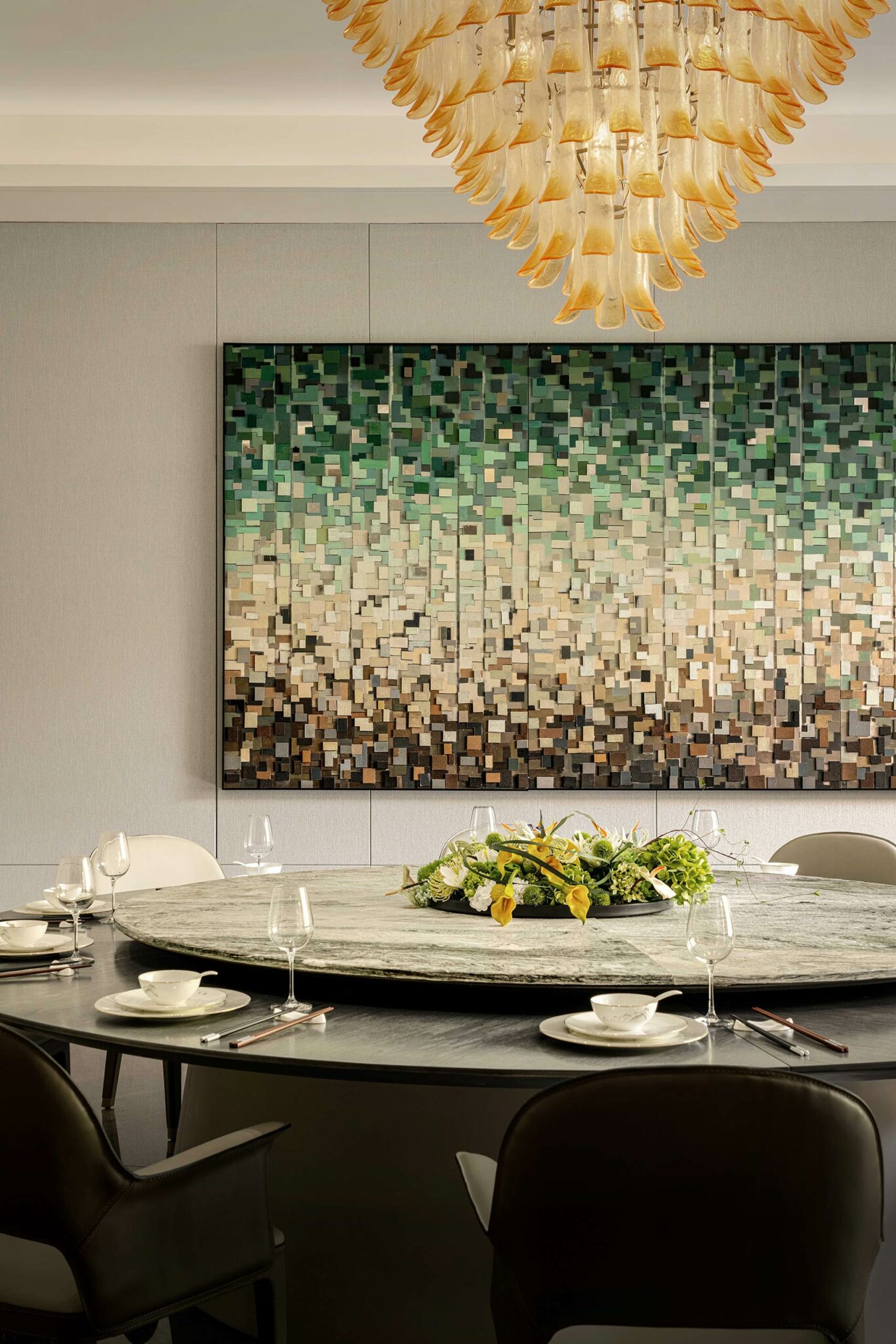 Elegant dining room with a large circular table set for six, beneath a golden chandelier. Abstract mosaic artwork in green and brown tones decorates the wall.