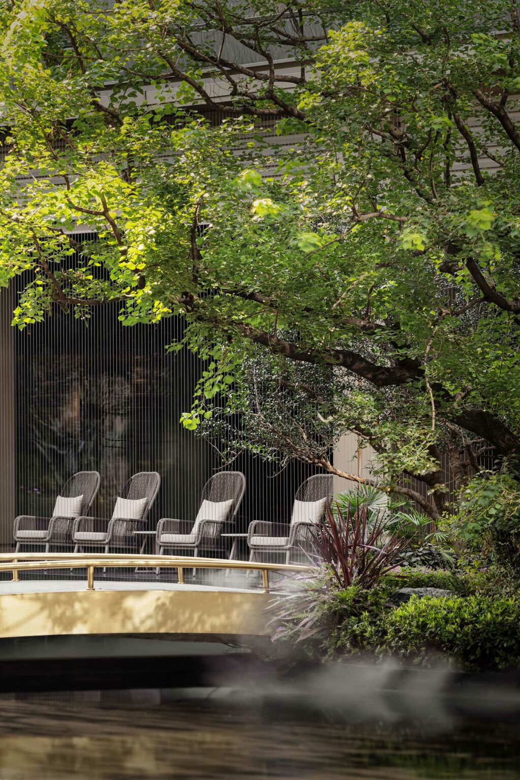 Four wicker chairs are lined up on a wooden bridge surrounded by lush green trees and shrubs, creating a serene and peaceful outdoor setting.