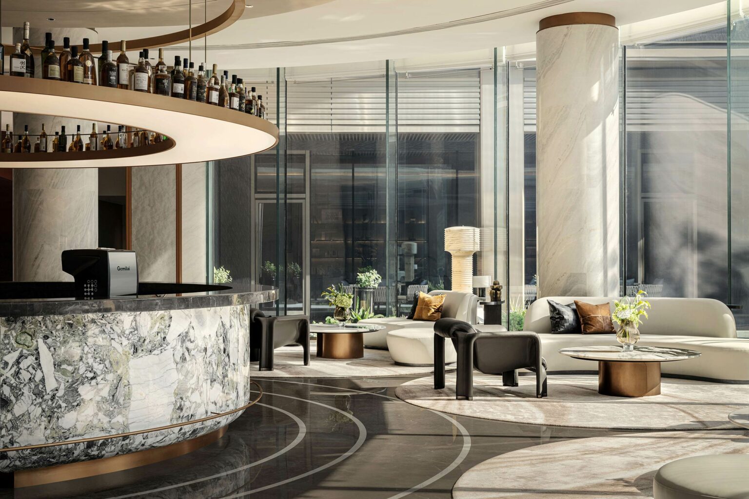 Modern hotel lobby with marble and glass décor. Circular bar with liquor bottles above. Elegant seating, soft natural light streaming in, calm atmosphere.
