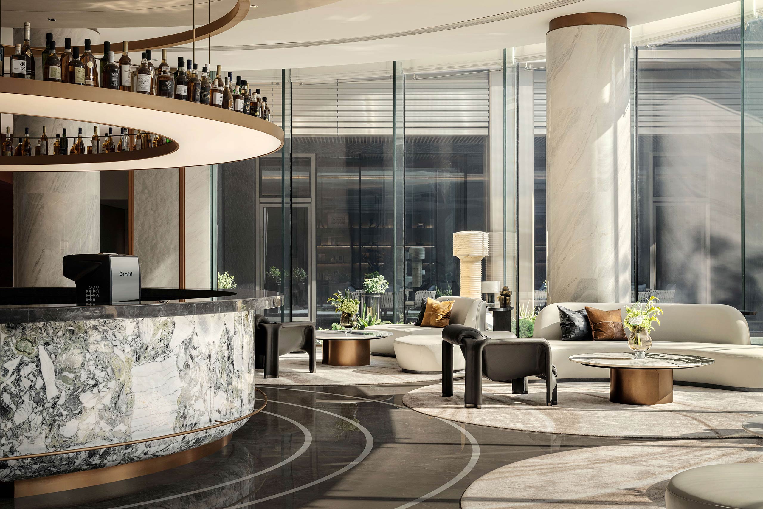 Modern hotel lobby with marble and glass décor. Circular bar with liquor bottles above. Elegant seating, soft natural light streaming in, calm atmosphere.