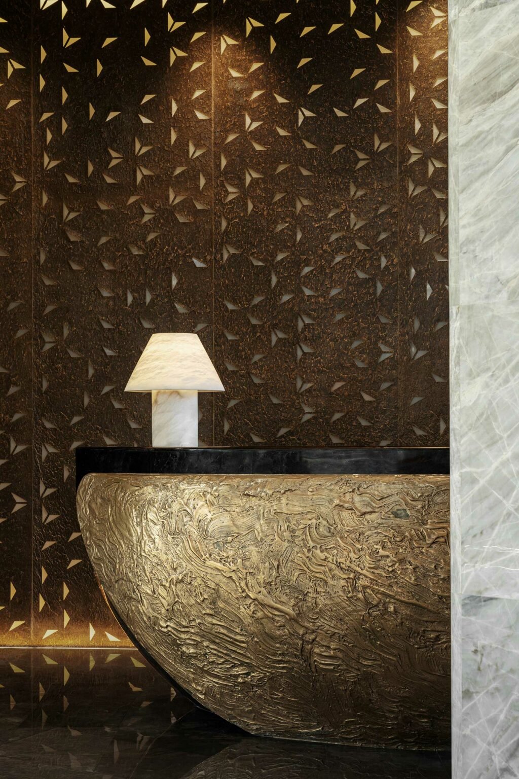 Intricately designed reception desk with a textured, gold surface, black trim, and a marble lamp. The background features a patterned, brown wall.