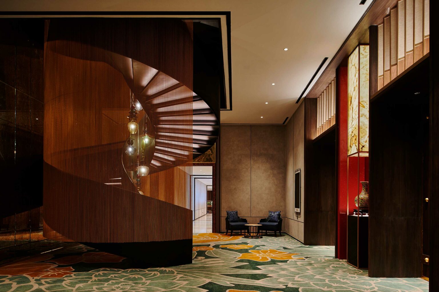 Elegant hotel lobby with a spiral staircase and hanging lights. The decor includes warm brown tones, patterned carpet, and two blue chairs. Serene atmosphere.