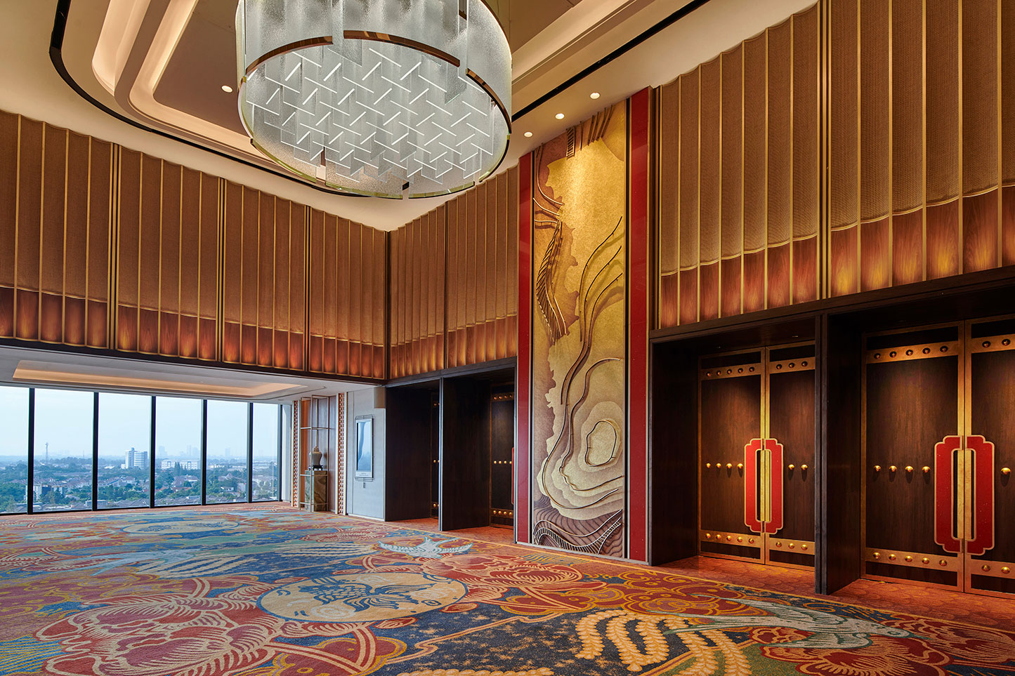 Luxurious hotel lift lobby with ornate patterned carpet and large abstract wall art. A geometric chandelier hangs from the ceiling. Large windows reveal a cityscape.