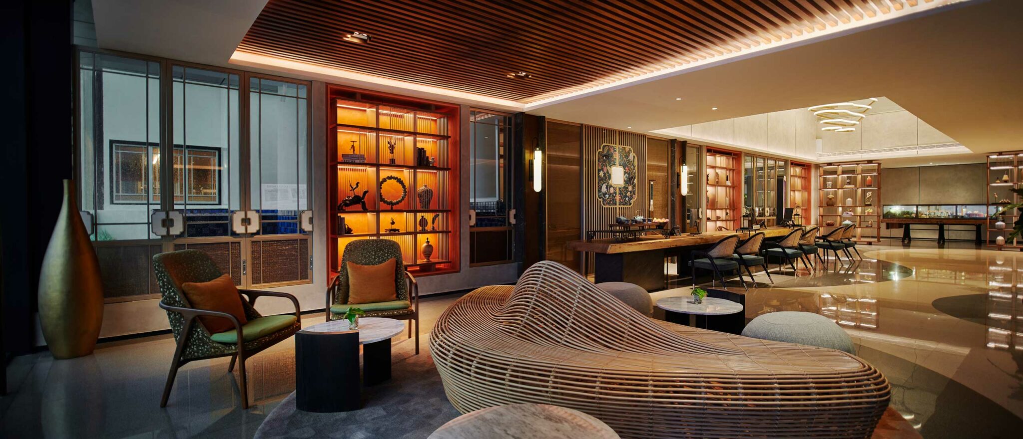 Modern hotel lounge with curved wooden seating, elegant armchairs, and a backlit shelf displaying art. Warm lighting creates a cozy, sophisticated ambiance.