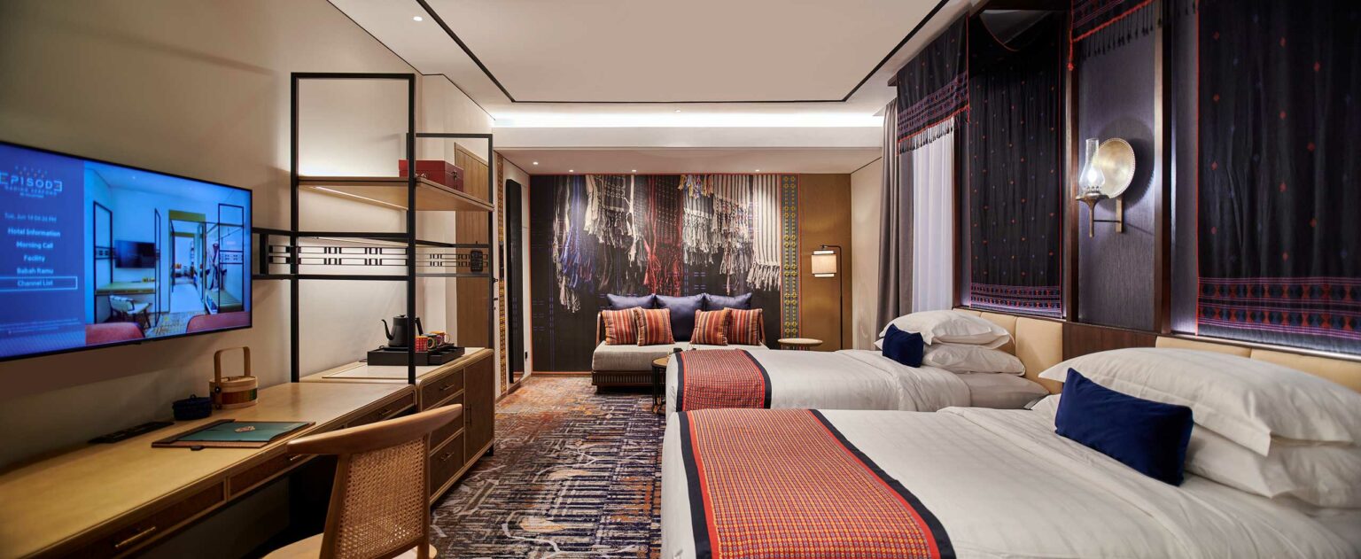 Modern hotel room with two neatly made beds featuring red and navy accents. A large TV is mounted on the wall, and a cozy seating area is in the corner, creating a welcoming and stylish ambiance.
