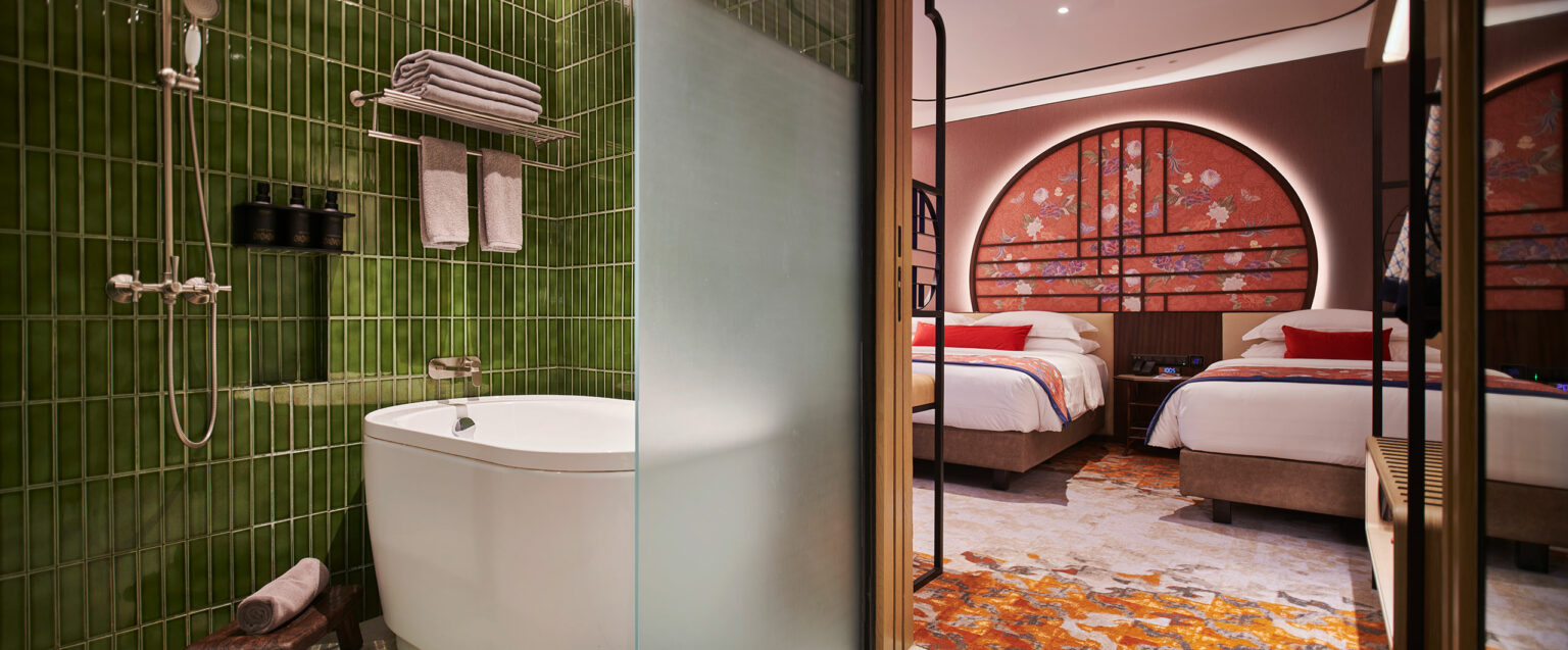 Elegant hotel room with a white tub against green tiled wall, towels, and toiletries. Nearby, two beds face a large, decorative floral headboard. Cozy and modern atmosphere.