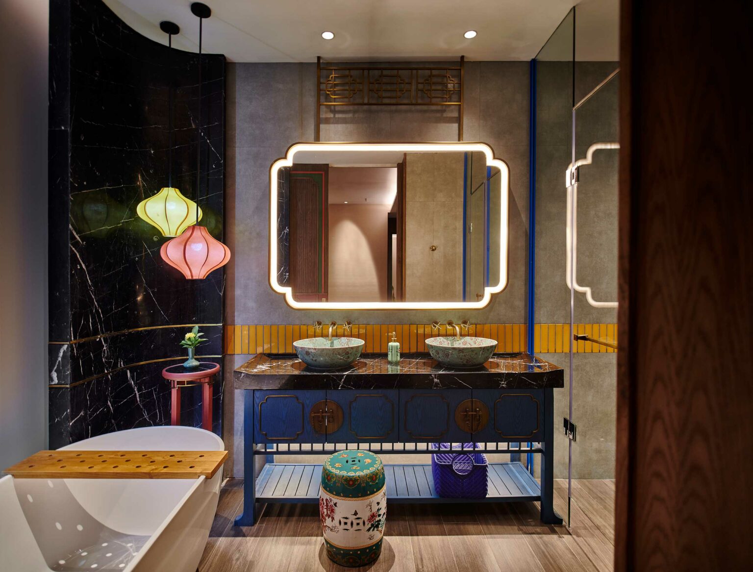Stylish guest room bathroom with illuminated mirror, colorful lanterns, twin vessel sinks on dark marble counter, and a decorative ceramic stool, conveying elegance.