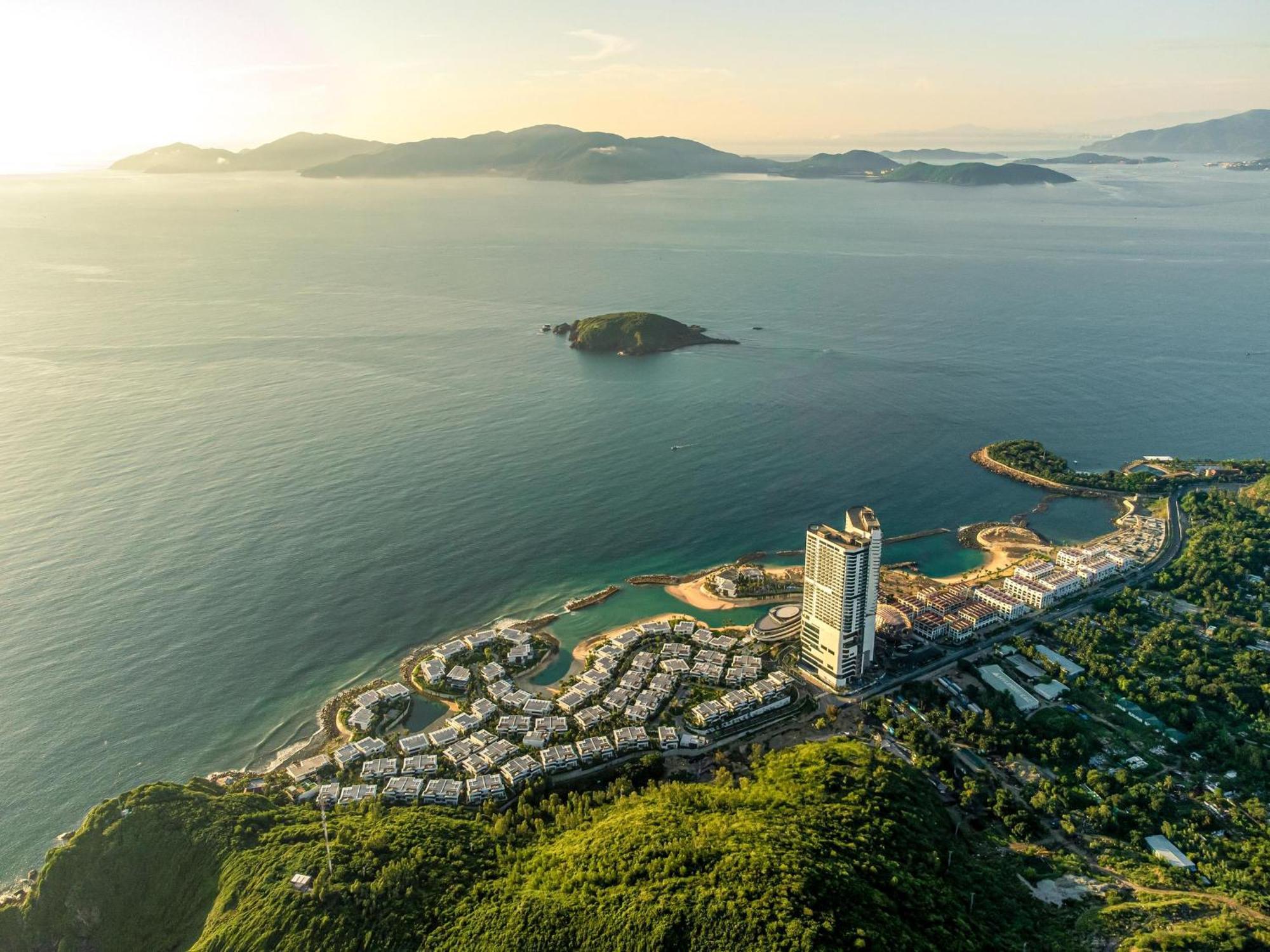 Aerial view of a coastal cityscape with a prominent tall building near the shore, surrounded by modern housing, lush green hills, and calm sea waters. The morning light casts a serene glow.