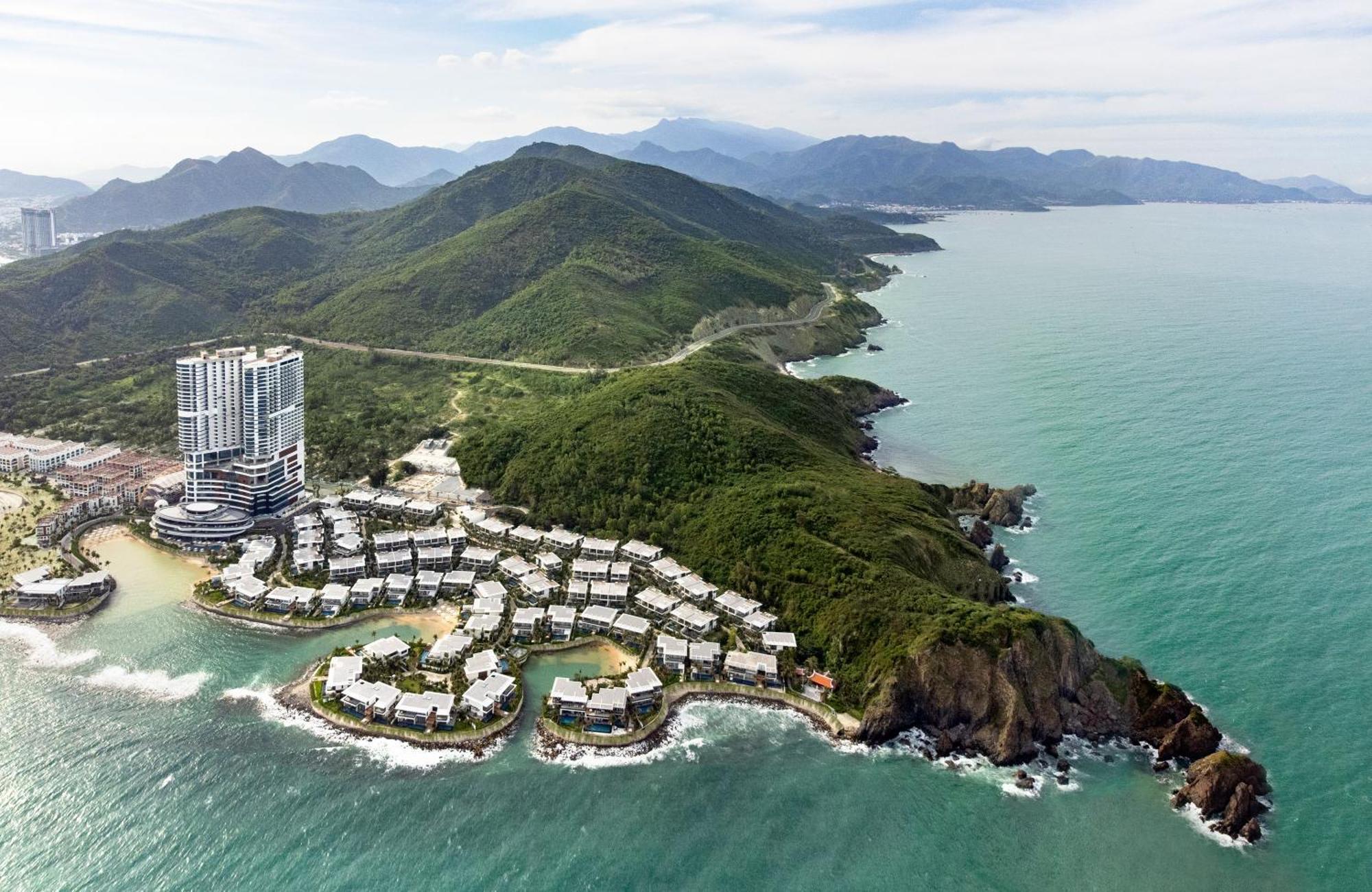 Aerial view of a coastal resort with white buildings nestled among lush green hills. The ocean waves crash against the rocky shoreline. Serene and picturesque.