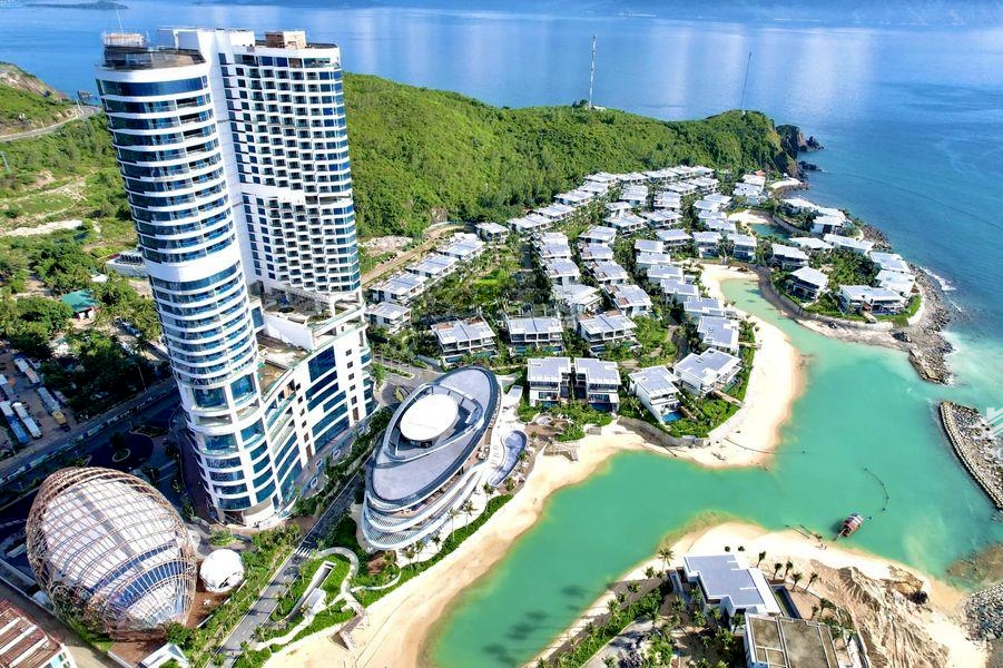 Aerial view of a luxurious coastal resort with two tall, modern cylindrical buildings, numerous white villas nestled amidst greenery, and turquoise waters.