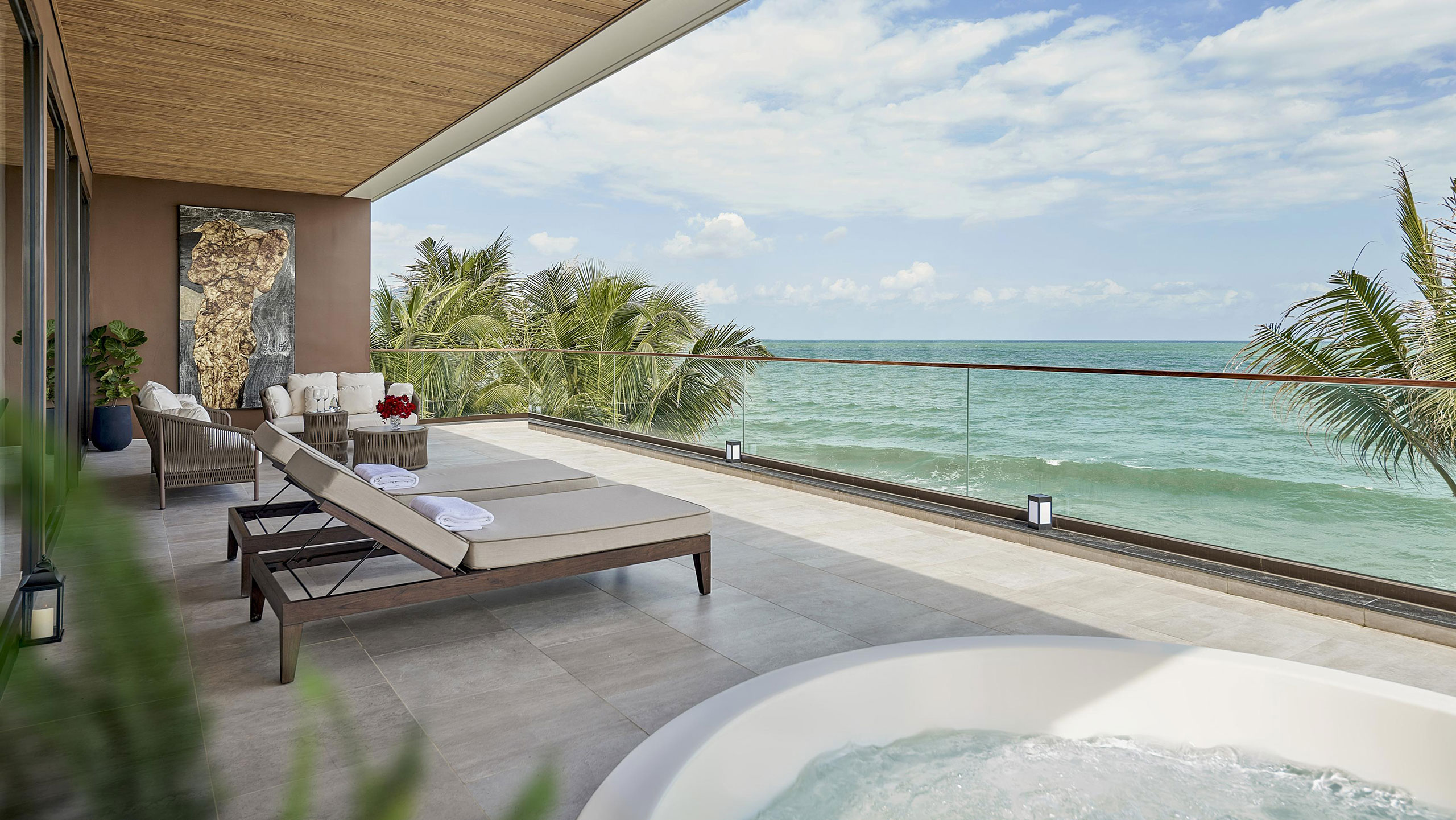 Spacious resort oceanfront balcony with two loungers, spa tub, art piece, and palm trees. Glass railing reveals serene sea and blue sky. Relaxing ambiance.