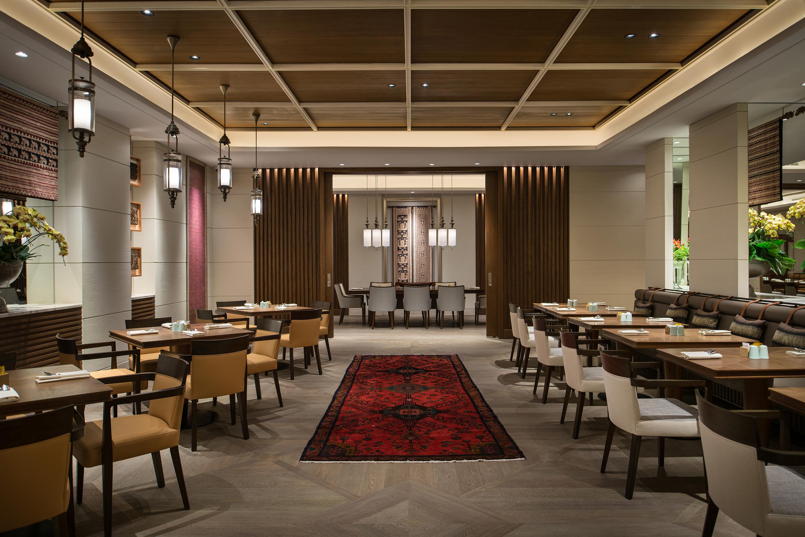 Luxurious restaurant interior with wooden ceiling, pendant lights, and a red Persian rug. Tables and chairs line the walls, creating a warm, elegant atmosphere.