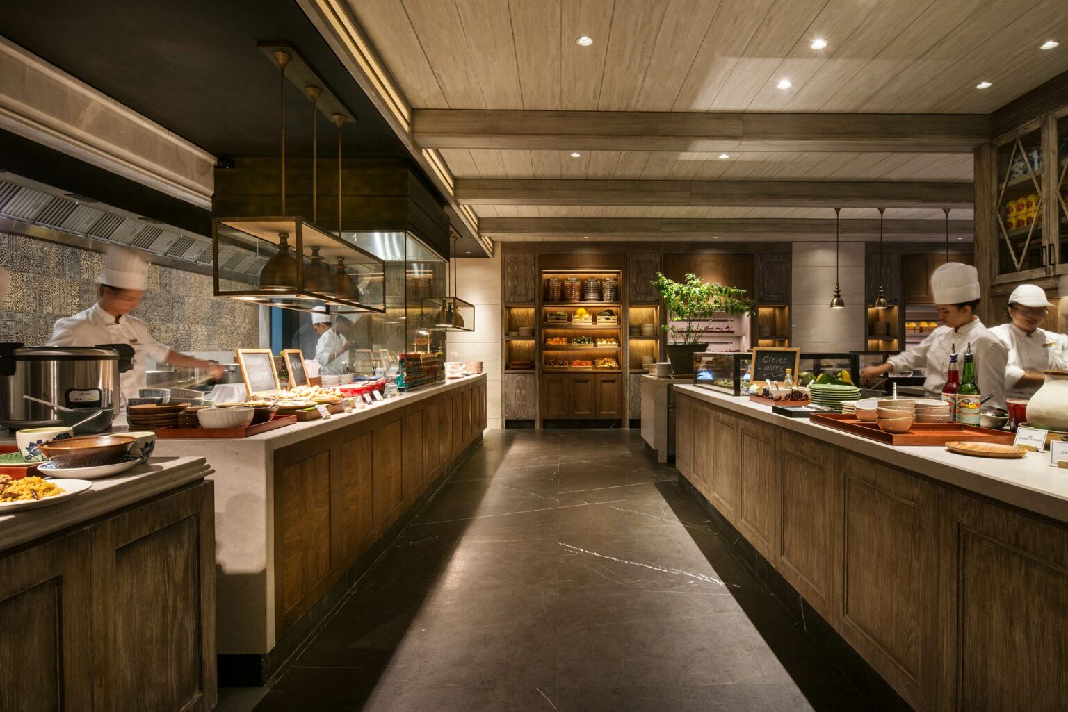 A spacious restaurant kitchen showcases chefs in white uniforms and hats preparing dishes. The warm lighting and wood accents create an inviting atmosphere.