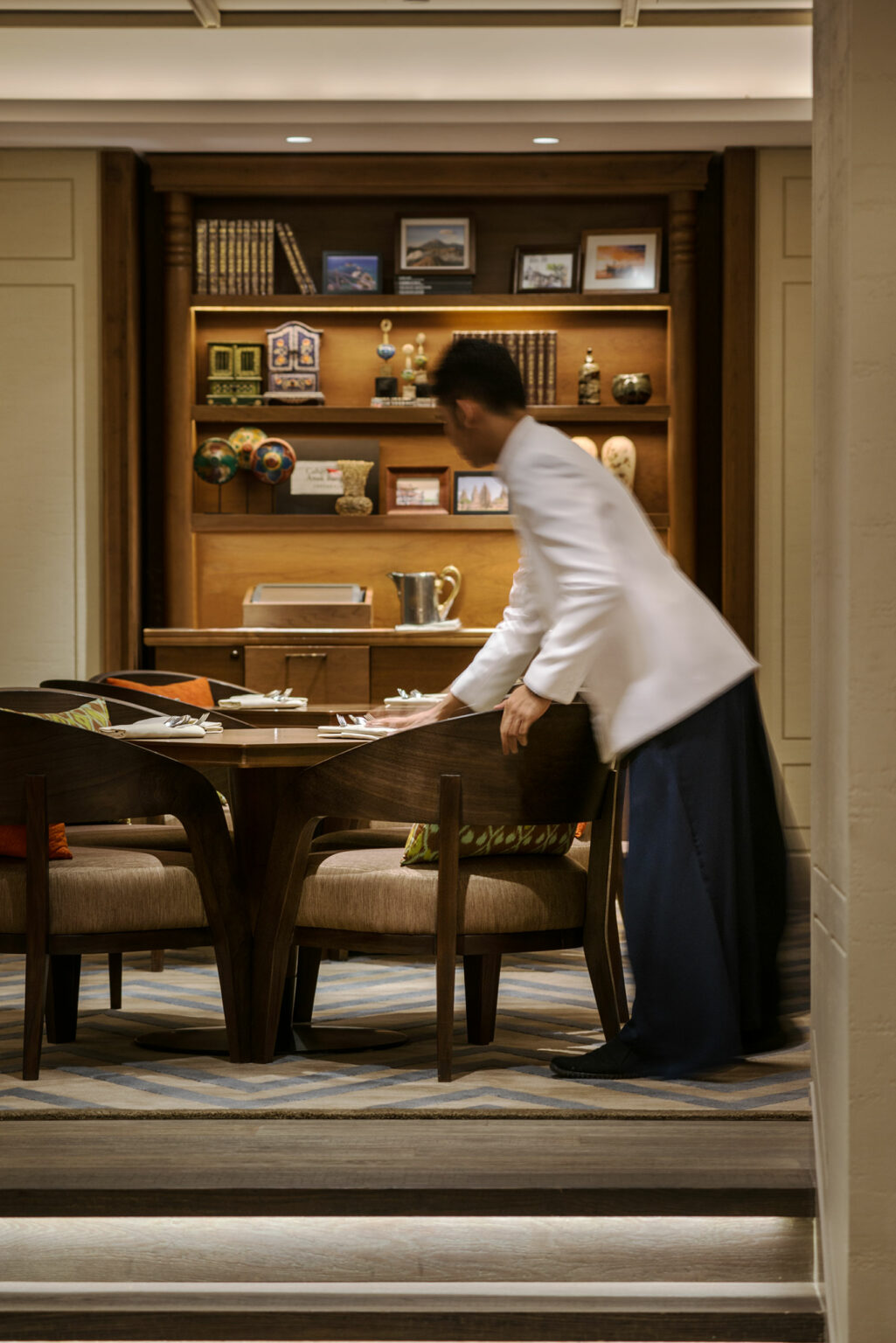 A waiter in a white jacket arranges chairs at an elegant dining table with a wooden bookshelf in the background, creating an upscale ambiance.