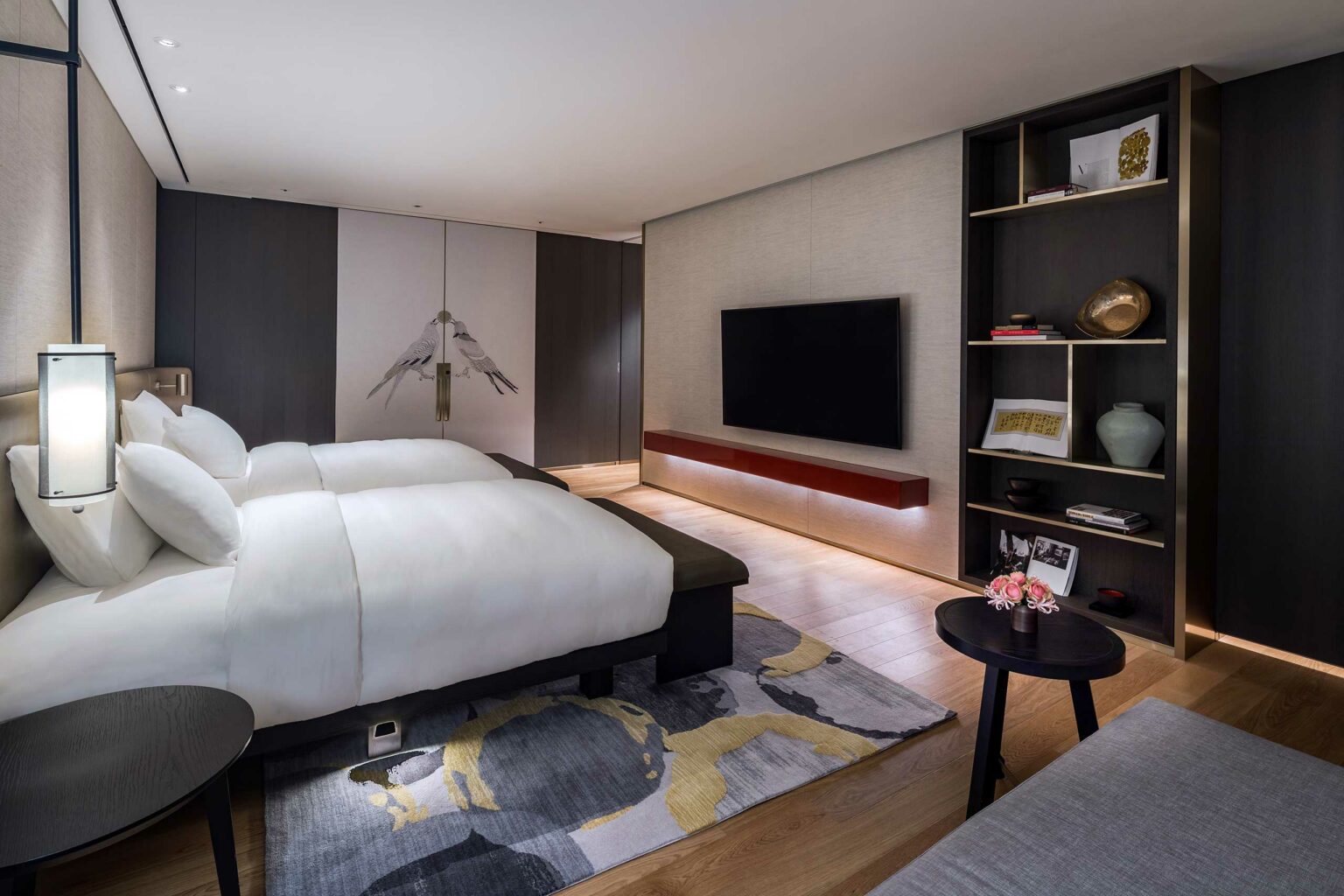 Modern hotel room with a plush white bed, dark wood accents, large TV, artistic wallpaper, and an elegant shelf. Warm lighting creates a cozy ambiance.